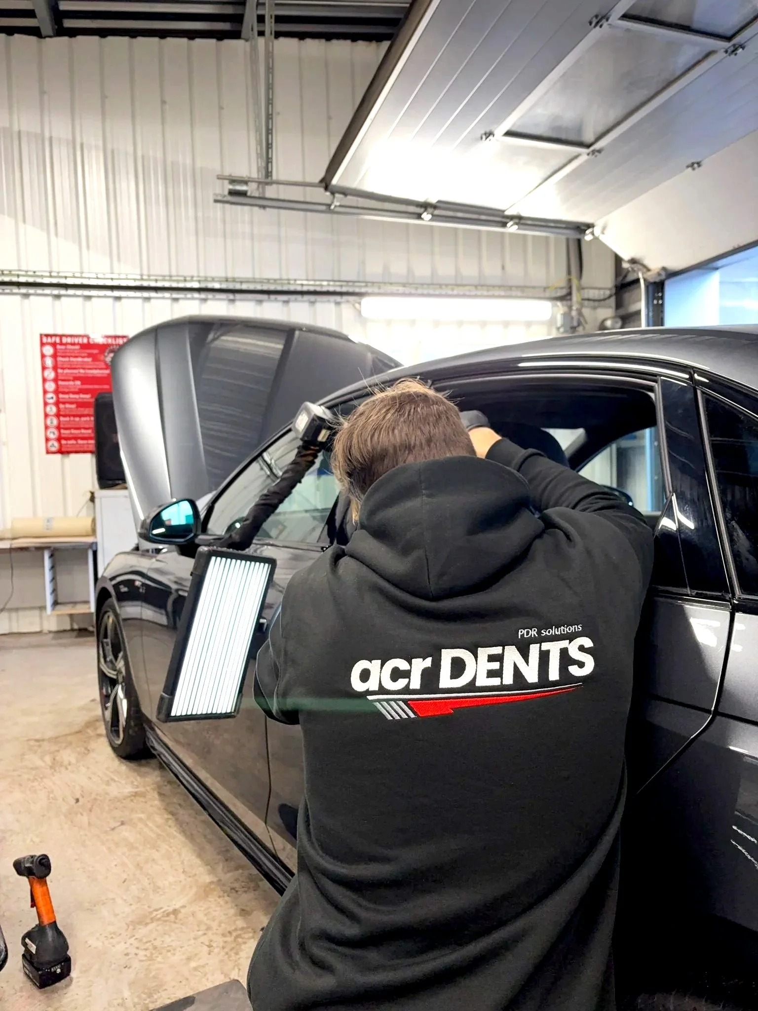 A mechanic working on a black car inside a garage with an open hood and tools nearby.