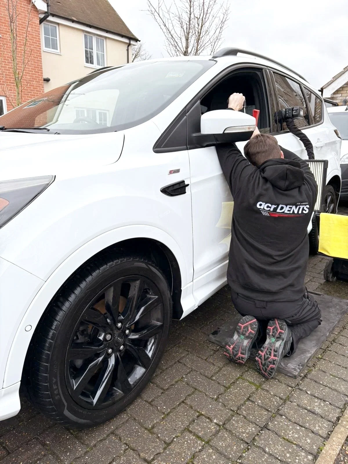 A man kneeling on a padded mat working on the front passenger side of a white SUV, possibly doing car repairs or maintenance, on a paved driveway outside a house.