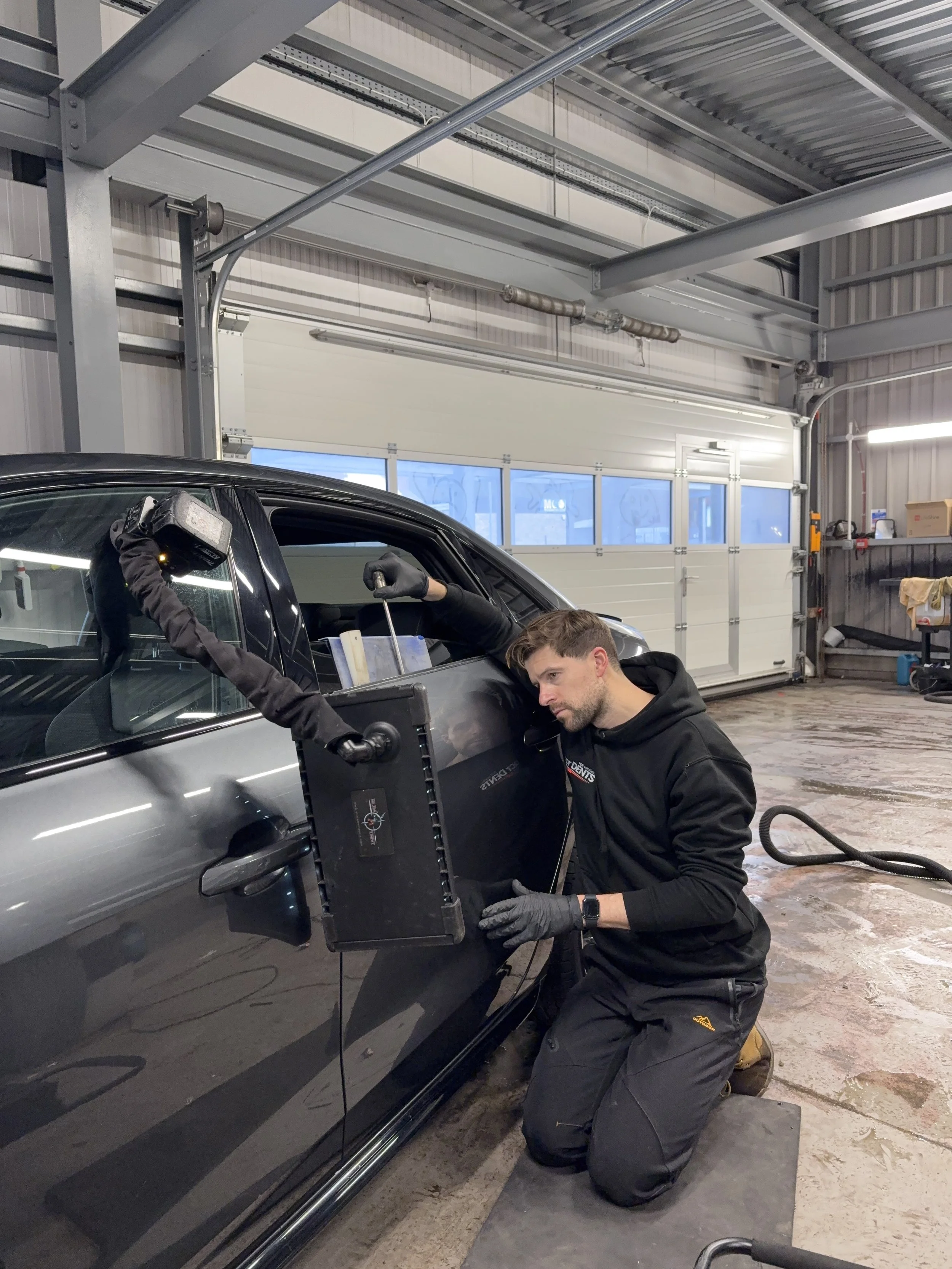 A man wearing black gloves and a black hoodie kneels outside a black car inside a garage, using specialized equipment on the vehicle's door frame.
