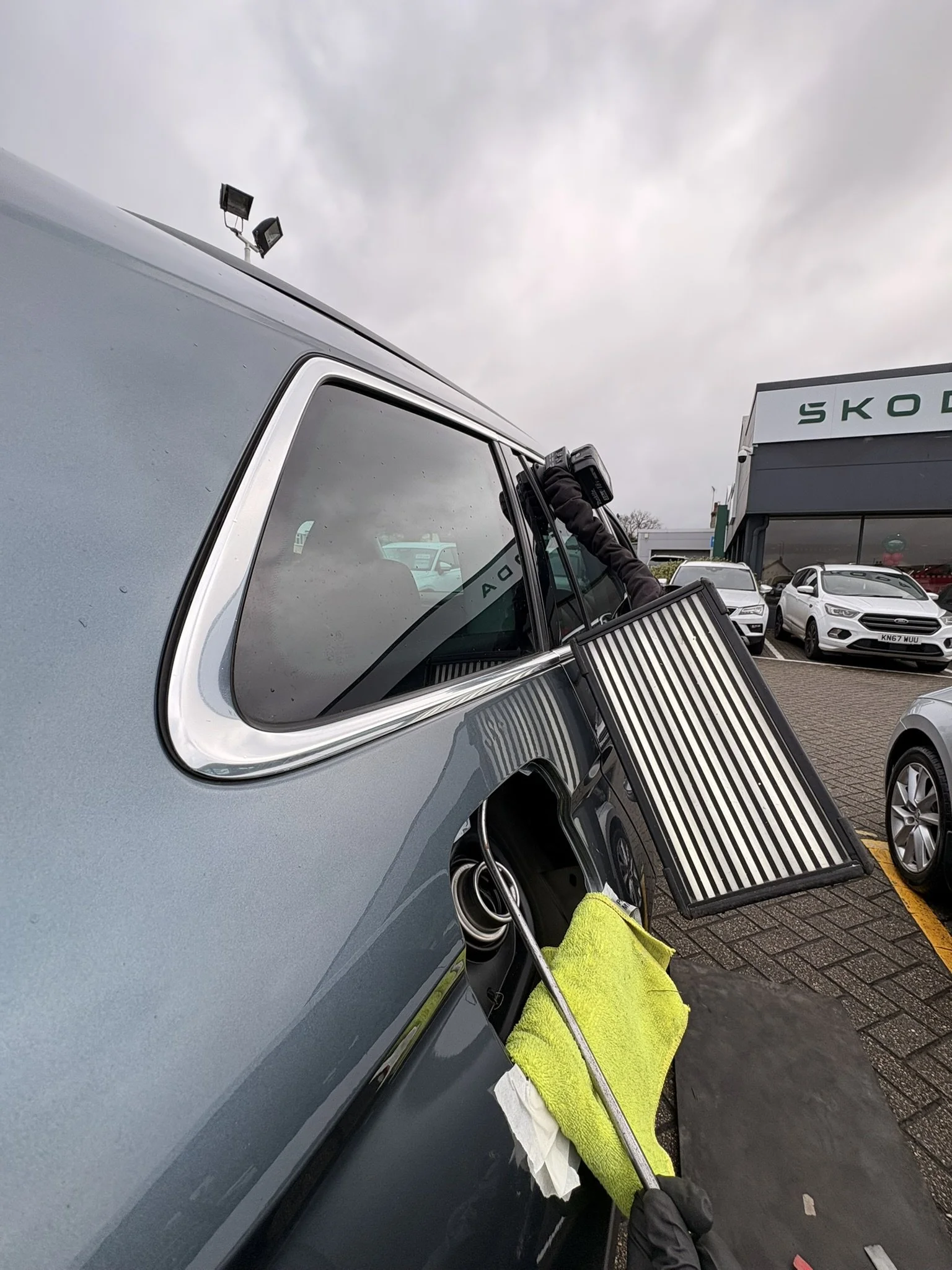 A person cleaning a vehicle's window with a squeegee, at a car dealership lot with multiple cars and a Skoda building in the background.