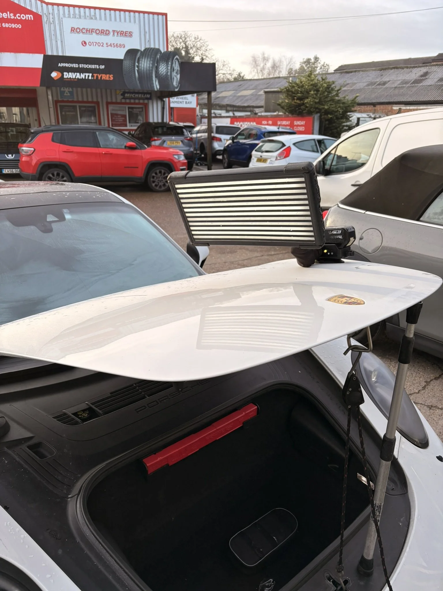 A white Porsche with a surfboard attached to its trunk and a portable light panel on top, parked in a car dealership lot with multiple other cars around and an automotive shop in the background.