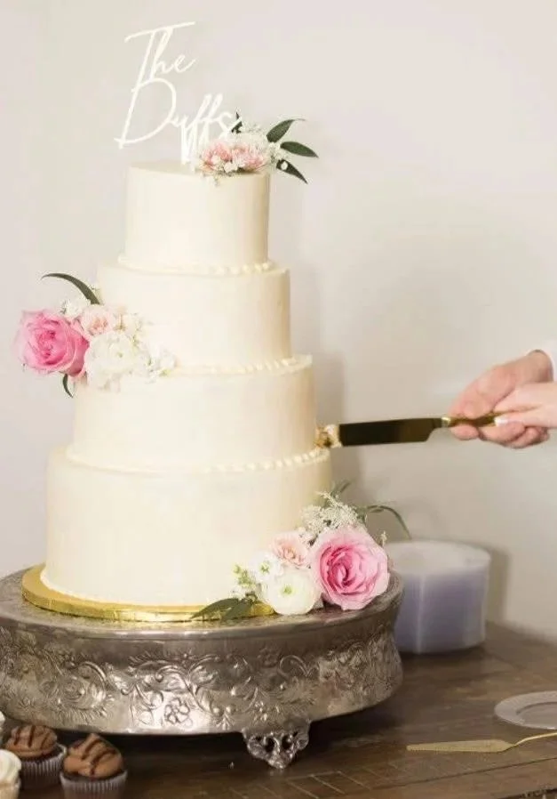 Four-tiered white wedding cake decorated with pink and white roses, floral accents, and a white script cake topper. A person is cutting the cake with a cake knife.