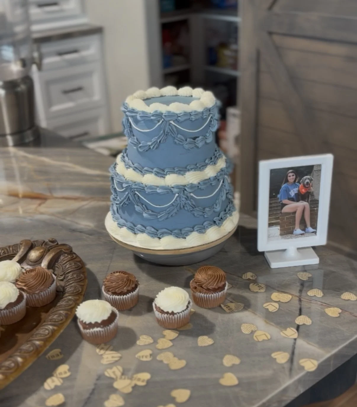 A two-tiered blue and white cake decorated with white frosting and blue icing swags, placed on a table. Cupcakes in white and brown wrappers with white and brown frosting are arranged nearby. A framed photograph of a young woman sitting on a brick le