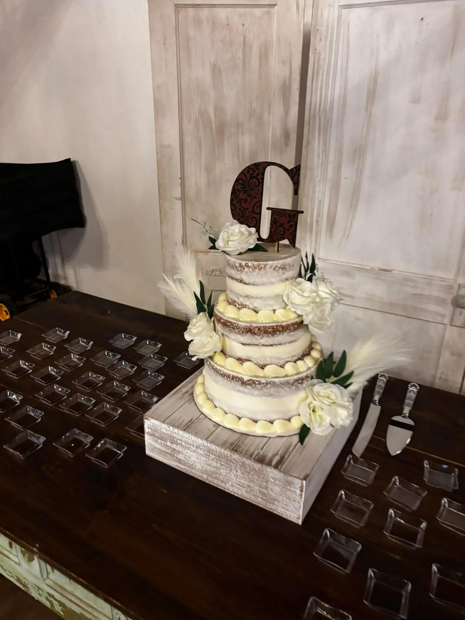 A three-tier naked wedding cake decorated with white flowers, greenery, and a large decorative letter 