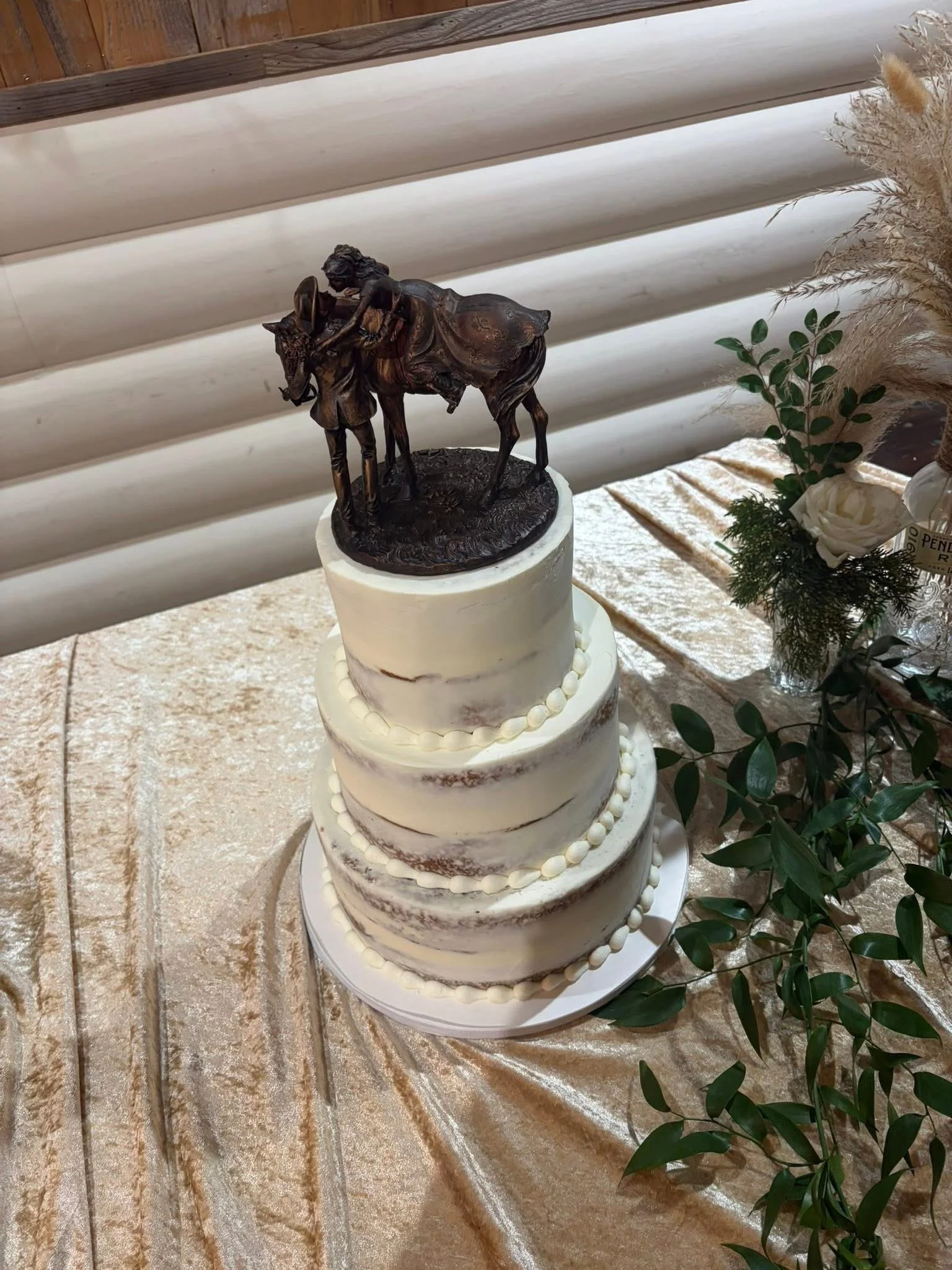 A three-tiered wedding cake with white icing and decorative border, topped with a bronze sculpture of a man on a donkey, and placed on a table with a gold satin tablecloth, surrounded by greenery and flowers.