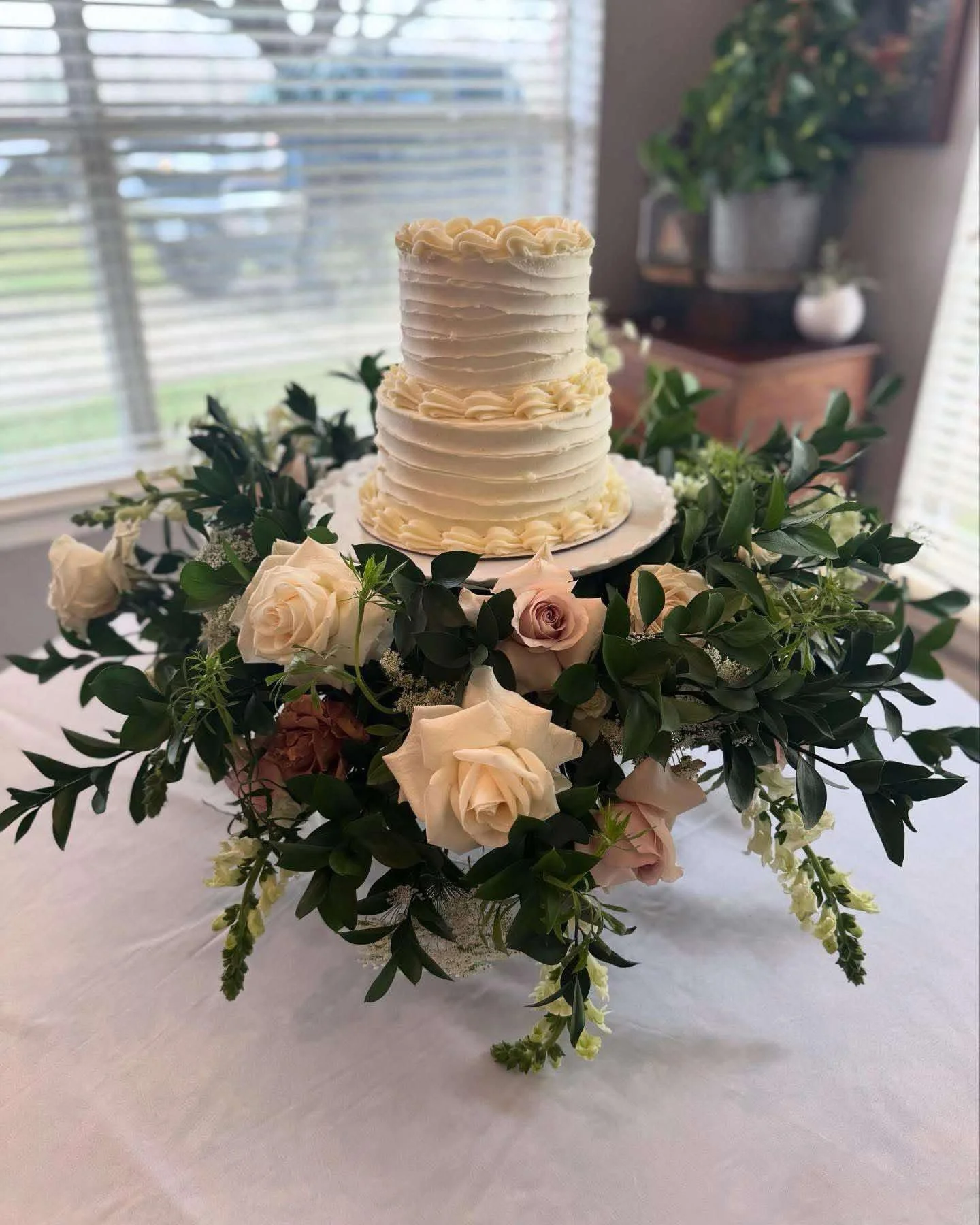 A wedding cake with two tiers, decorated with cream-colored frosting and floral accents, on a table surrounded by a floral arrangement of roses and greenery.