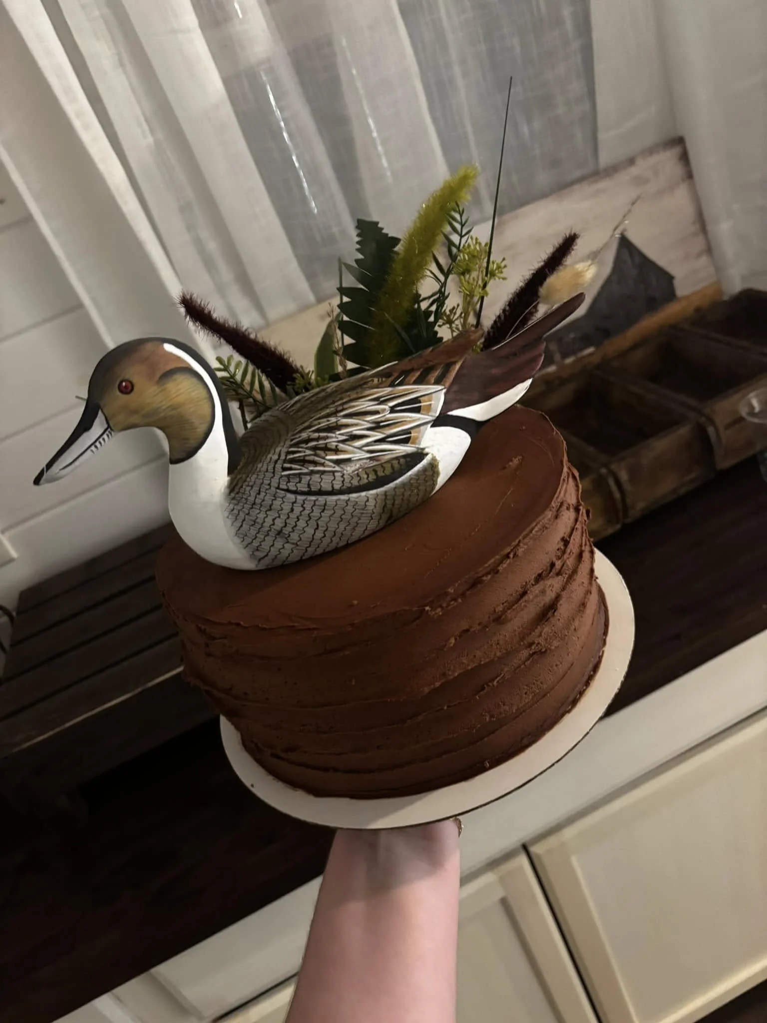 A decorative cake with a mounted duck decoy on top, surrounded by artificial greenery and feathers, set on a wooden surface in a cozy indoor setting.