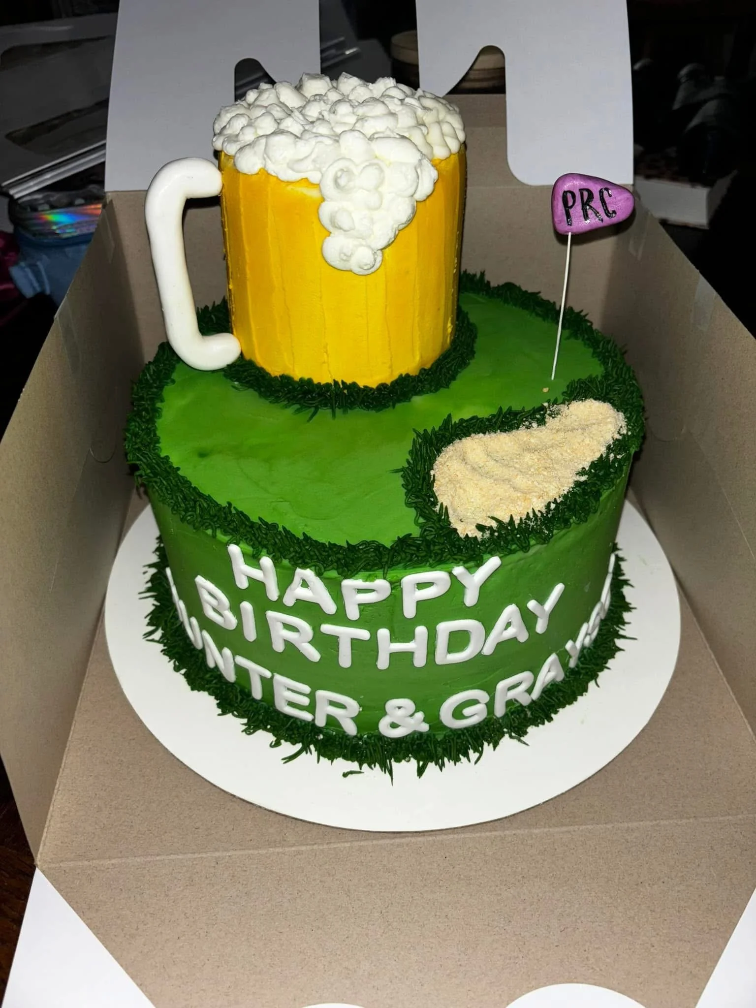 A birthday cake with a green exterior and a beer mug-shaped cake on top, decorated with white piping and a small pink flag that says 'PRC.'