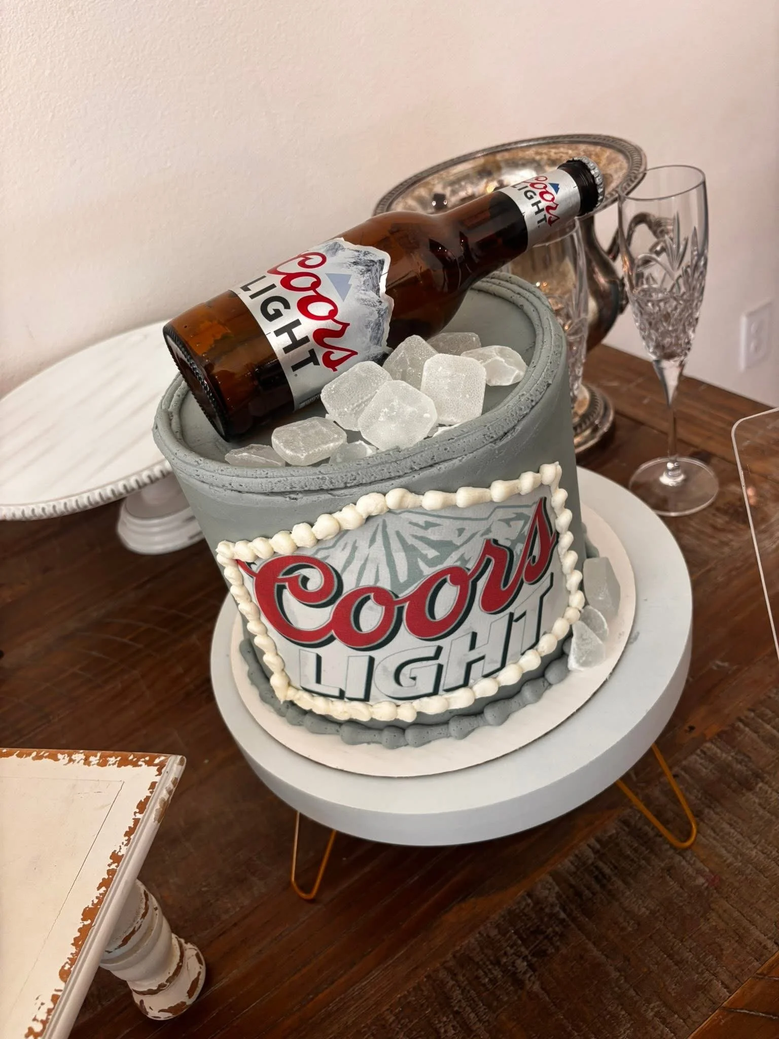 A Coors Light beer bottle on a display resembling an ice bucket, with ice cubes, placed on a white round table. A Coors Light branded cake with a graffiti-style design is also on the table. In the background, there is an ornate silver pitcher and a c