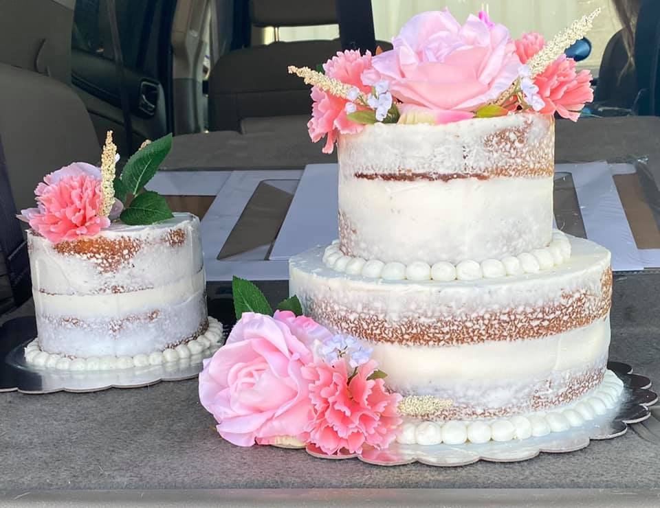 Two pink frosted wedding cakes with floral decorations on a gray table, one larger multi-tiered cake and one smaller single-tier cake, with watercolor-like pink flowers and green leaves.