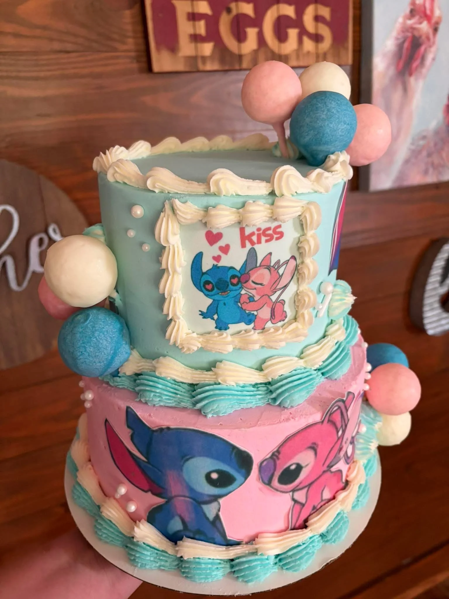 A two-tier birthday cake decorated with characters from the cartoon series 'Lilo & Stitch.' The top tier is light blue with cream-colored piping and contains a picture of Stitch and Angel with the word 'kiss' and red hearts. The bottom tier is pink w