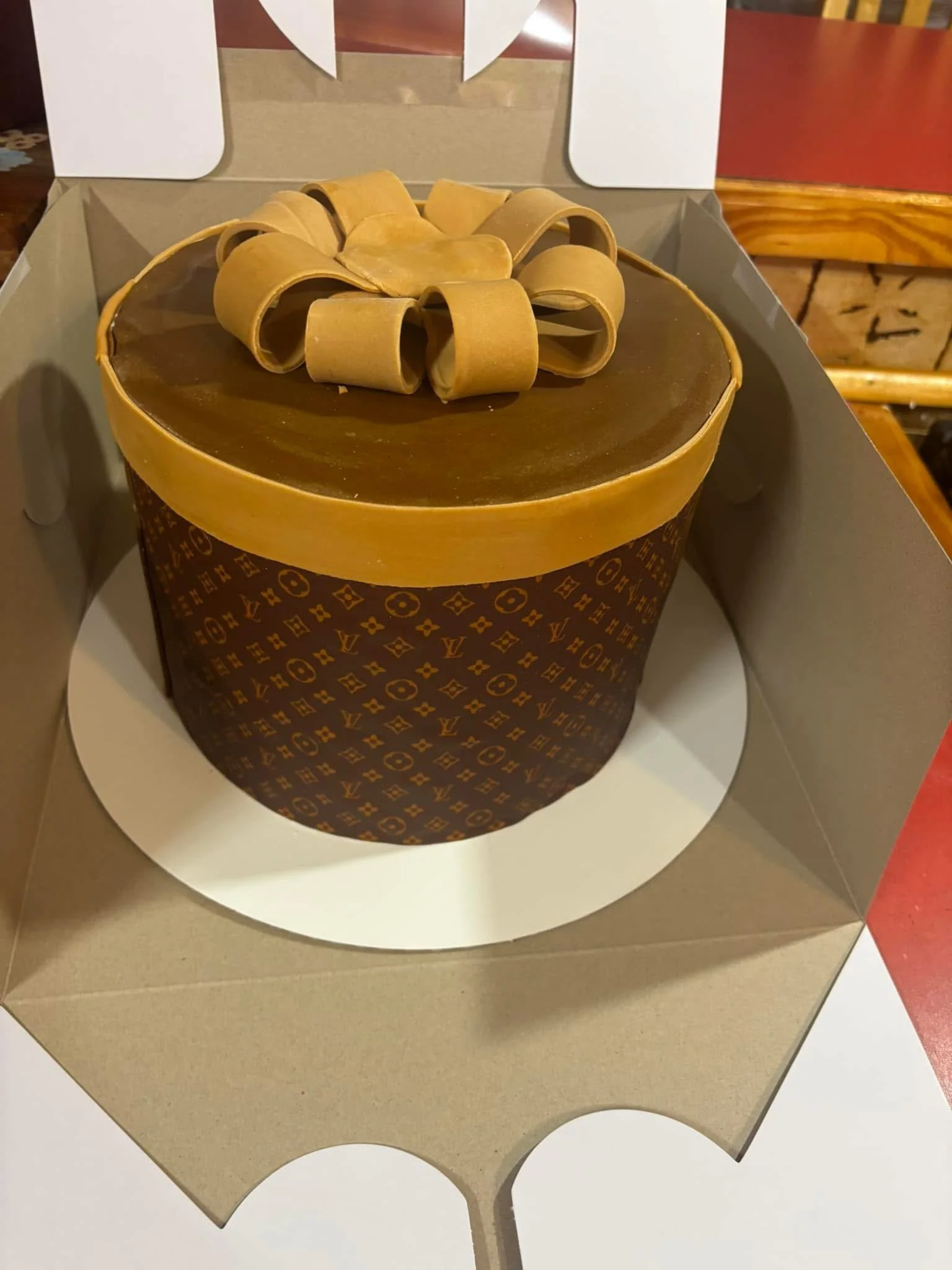 A round chocolate cake with a gold band around the top edge, decorated with a bow made of gold-colored ribbons on top, inside an open cardboard box.