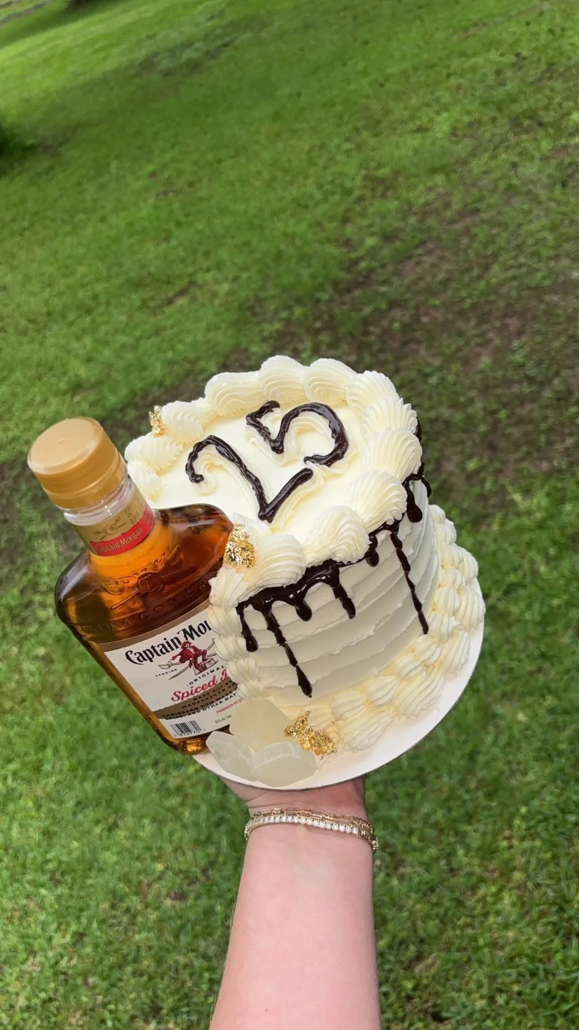 A hand holding a birthday cake with the number 25 written on top, decorated with white frosting, chocolate drizzle, and gold accents, with a small bottle of Captain Morgan Spiced Rum attached to the side of the cake.