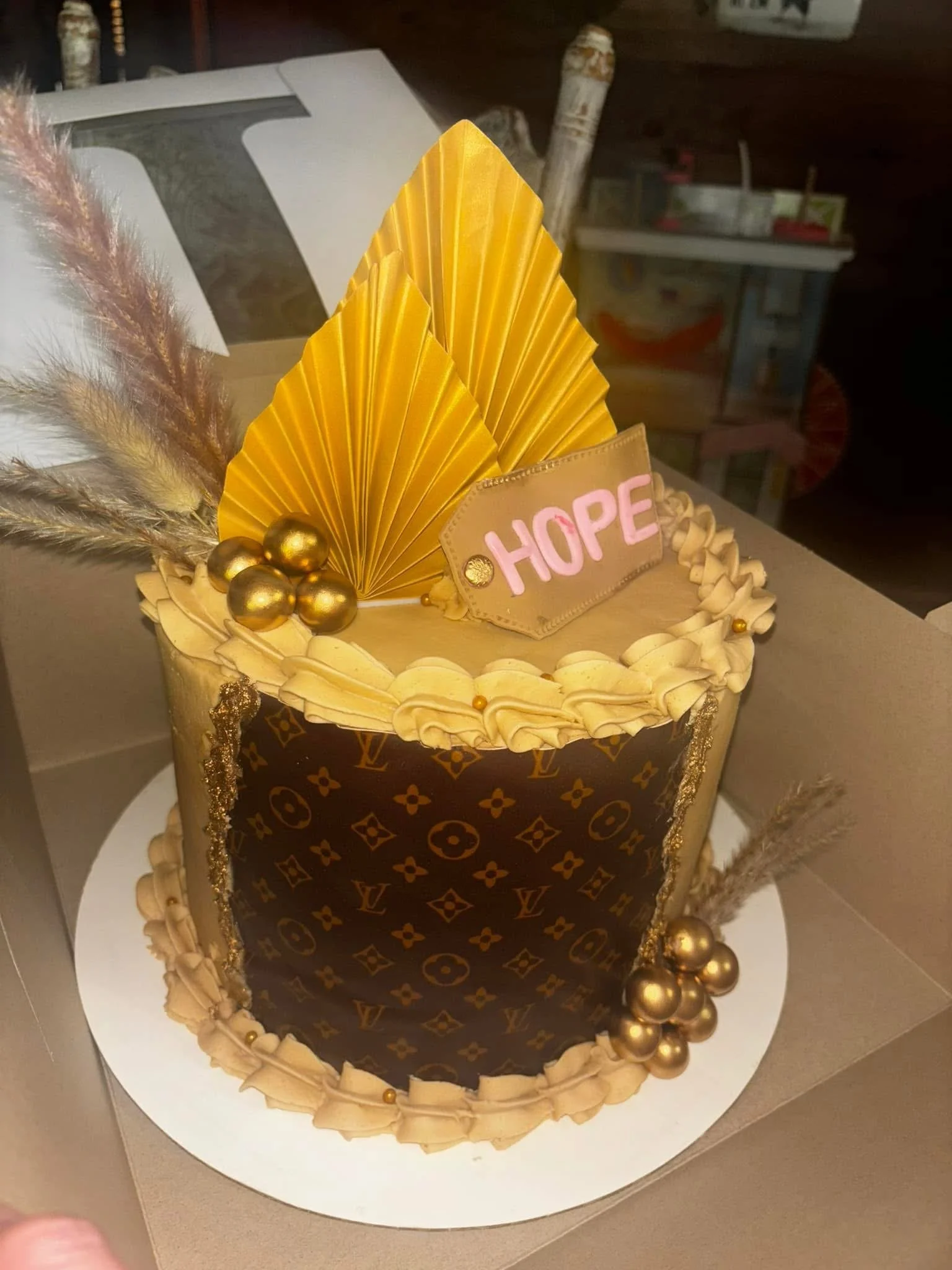 A luxurious cake decorated with gold leaves, gold berries, and feathers, with a Louis Vuitton pattern on the sides and a sign that says 'HOPE' on top.