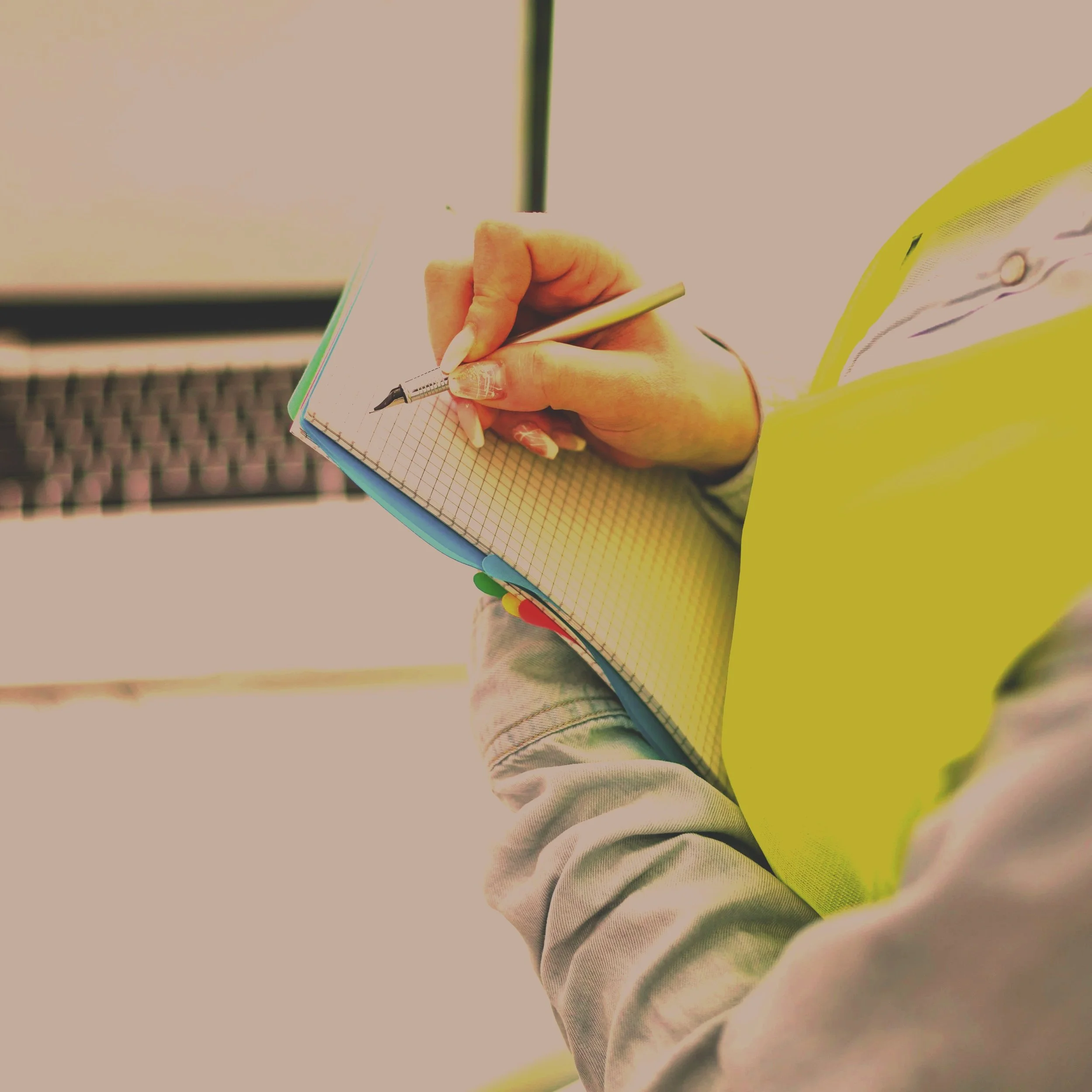 Close-up of a person holding a spiral notebook and pen, with their arm partially visible, wearing a yellow jacket, and a blurred background.