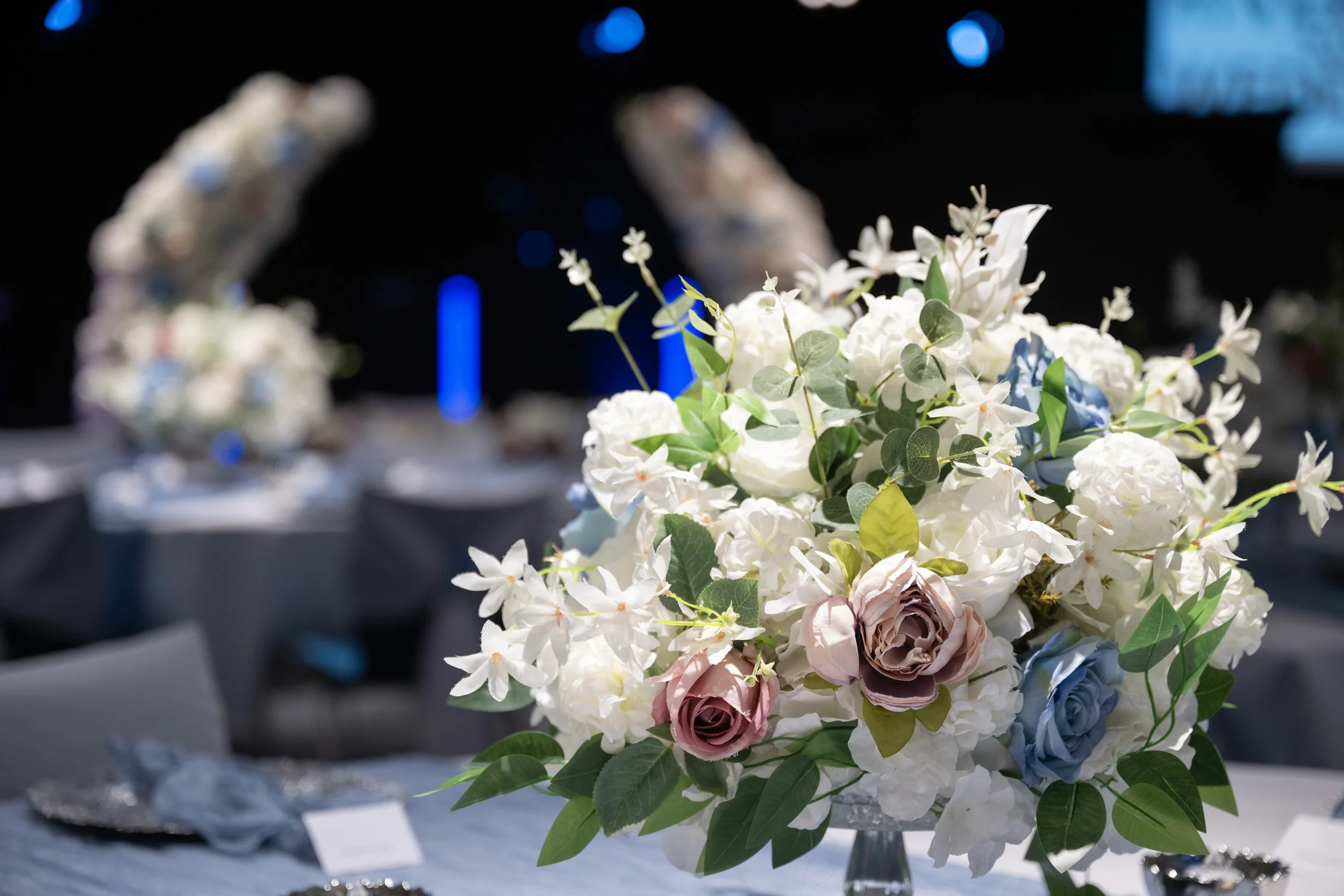 A large centerpiece floral arrangement with white, pink, and blue roses, white hydrangeas, white orchids, and green leaves on a table at an event.
