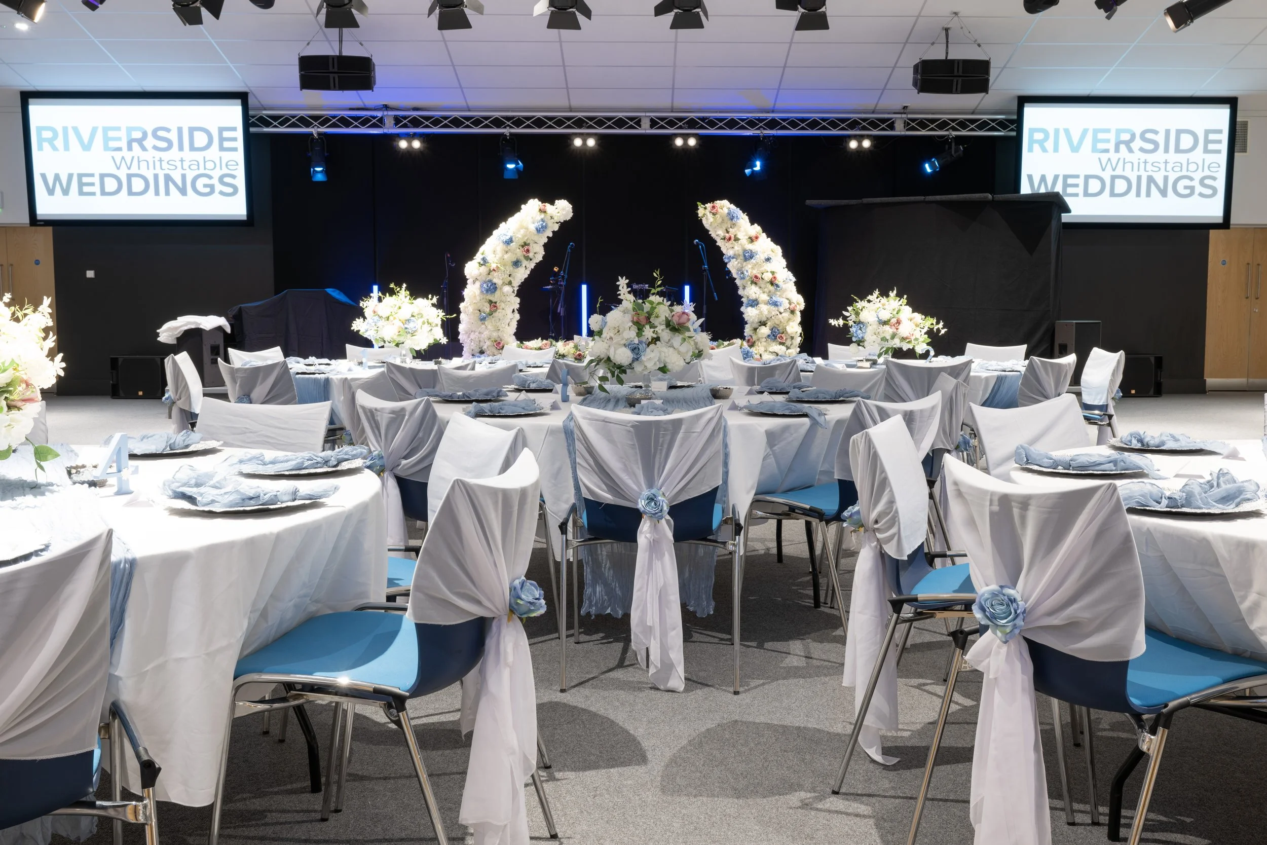 Wedding reception setup with decorated tables, white and blue accents, floral centerpieces, and a stage with a backdrop and screens displaying 'Riverside Whitstable Weddings'.
