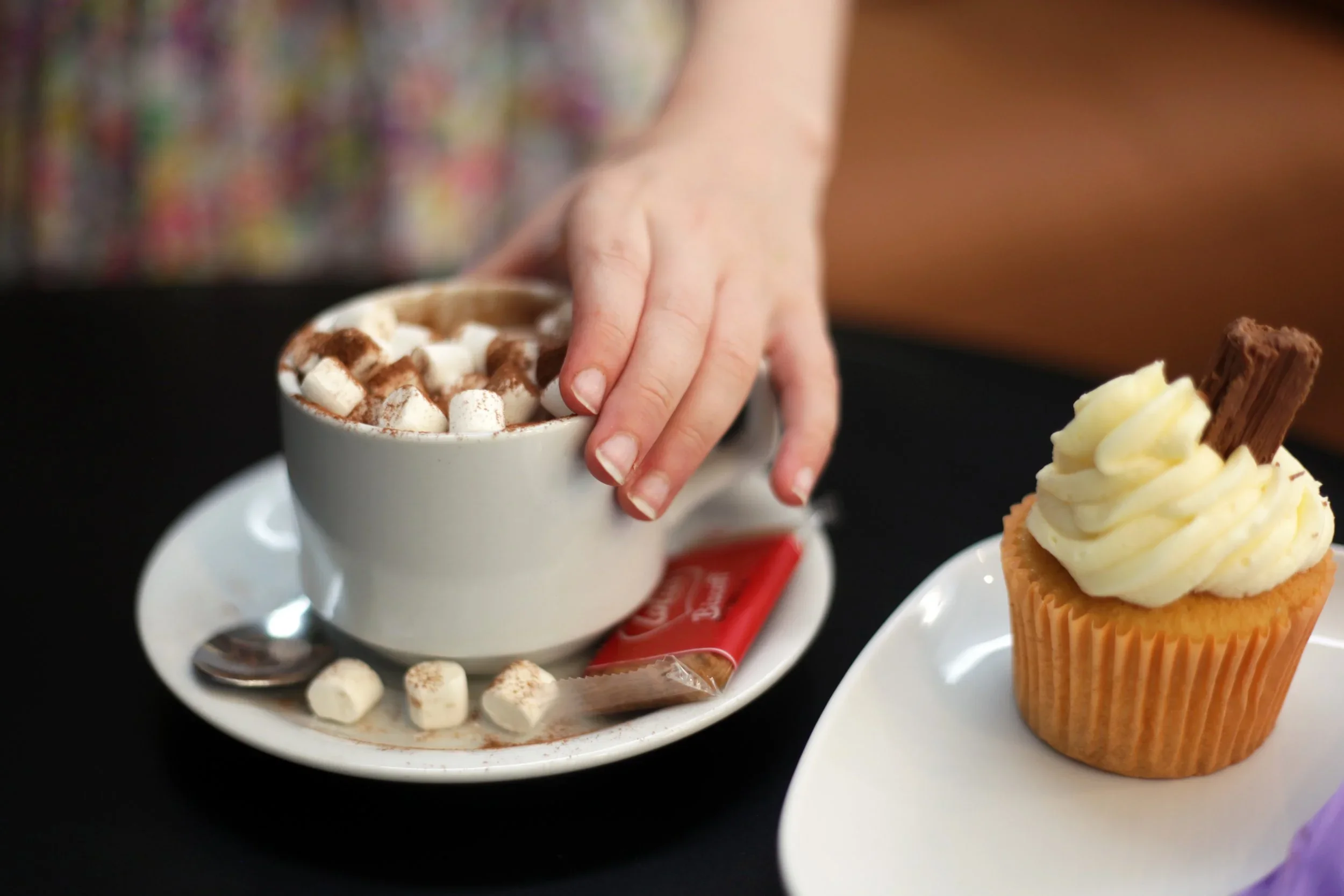 A person reaching for a cup of hot chocolate topped with mini marshmallows, next to a cupcake with white frosting and a chocolate wafer on top.
