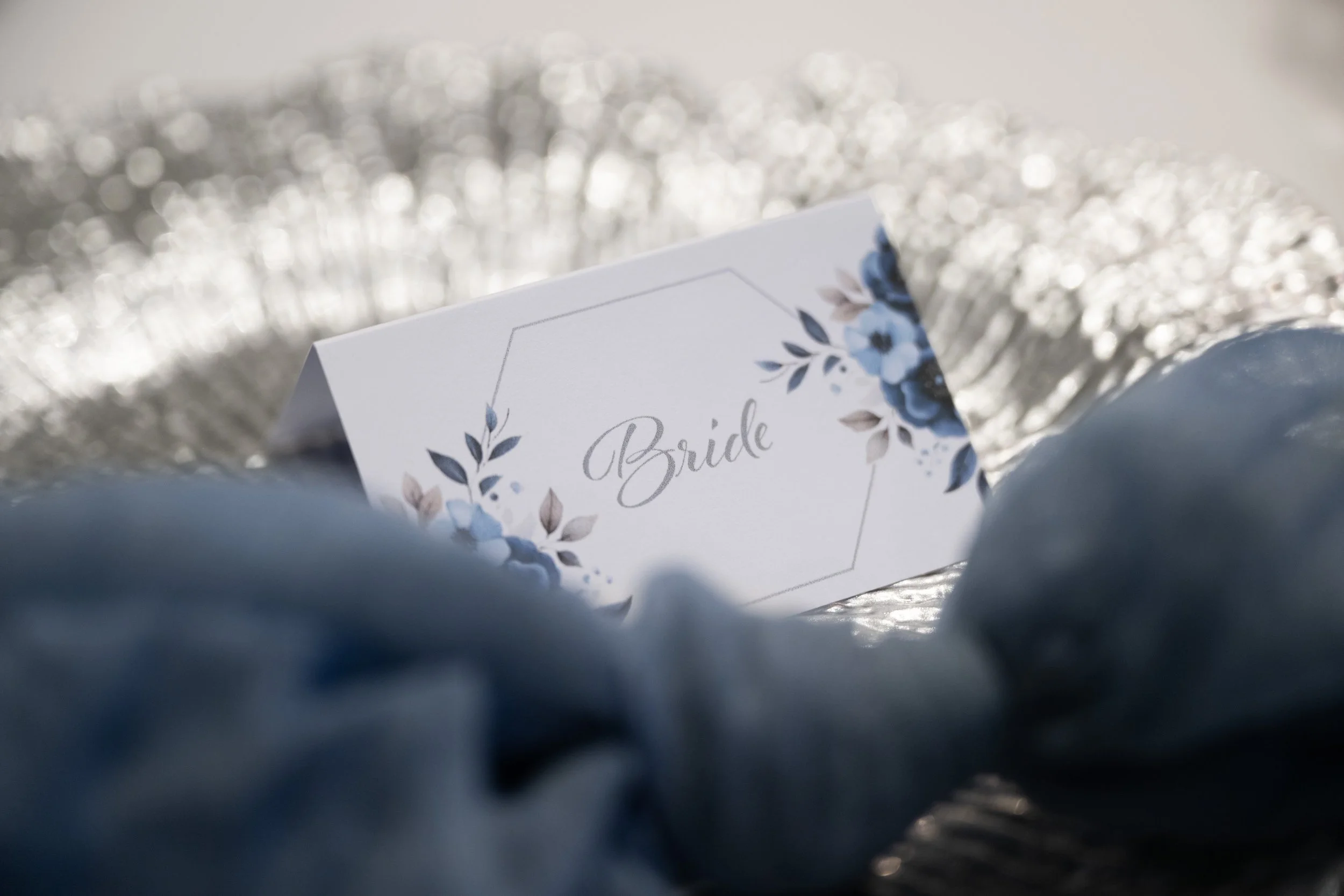 A white card with floral design and the word 'Bride' written on it, resting on silver textured fabric and partially surrounded by a soft blue cloth.