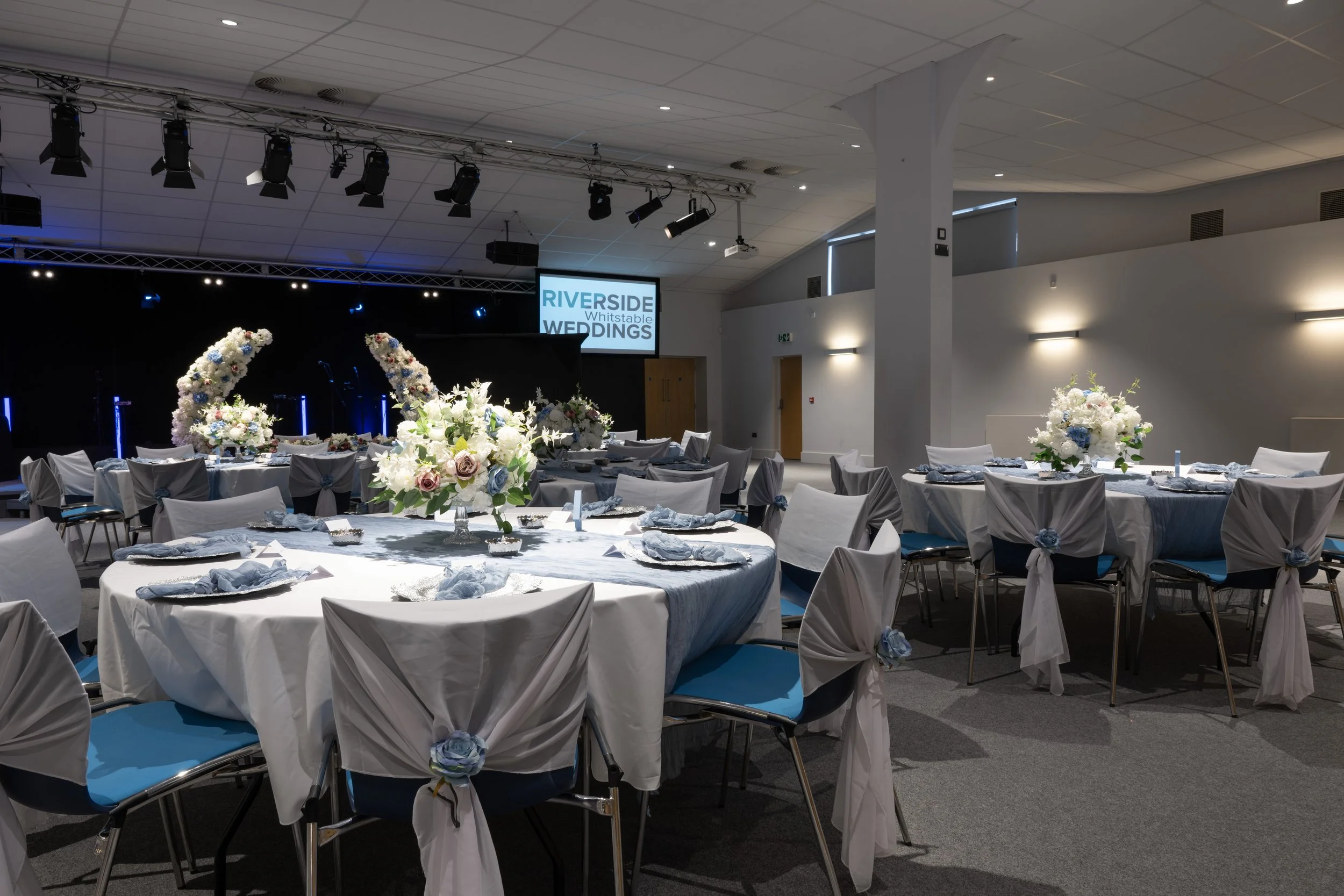 Elegant wedding reception hall with round tables decorated with white and pastel floral centerpieces, chairs covered with white fabric and blue accents, set for a meal, with a stage and screen displaying 'RIVERSIDE Whitstable WEDDINGS' in the backgro