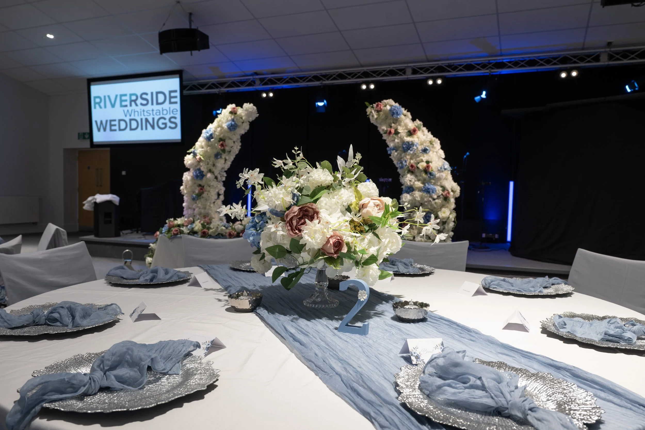 A decorated wedding reception table with a floral centerpiece, silver chargers with blue napkins, and a tall arch decorated with flowers in the background. A sign on the wall reads "RIVERSIDE Whistable WEDDINGS".