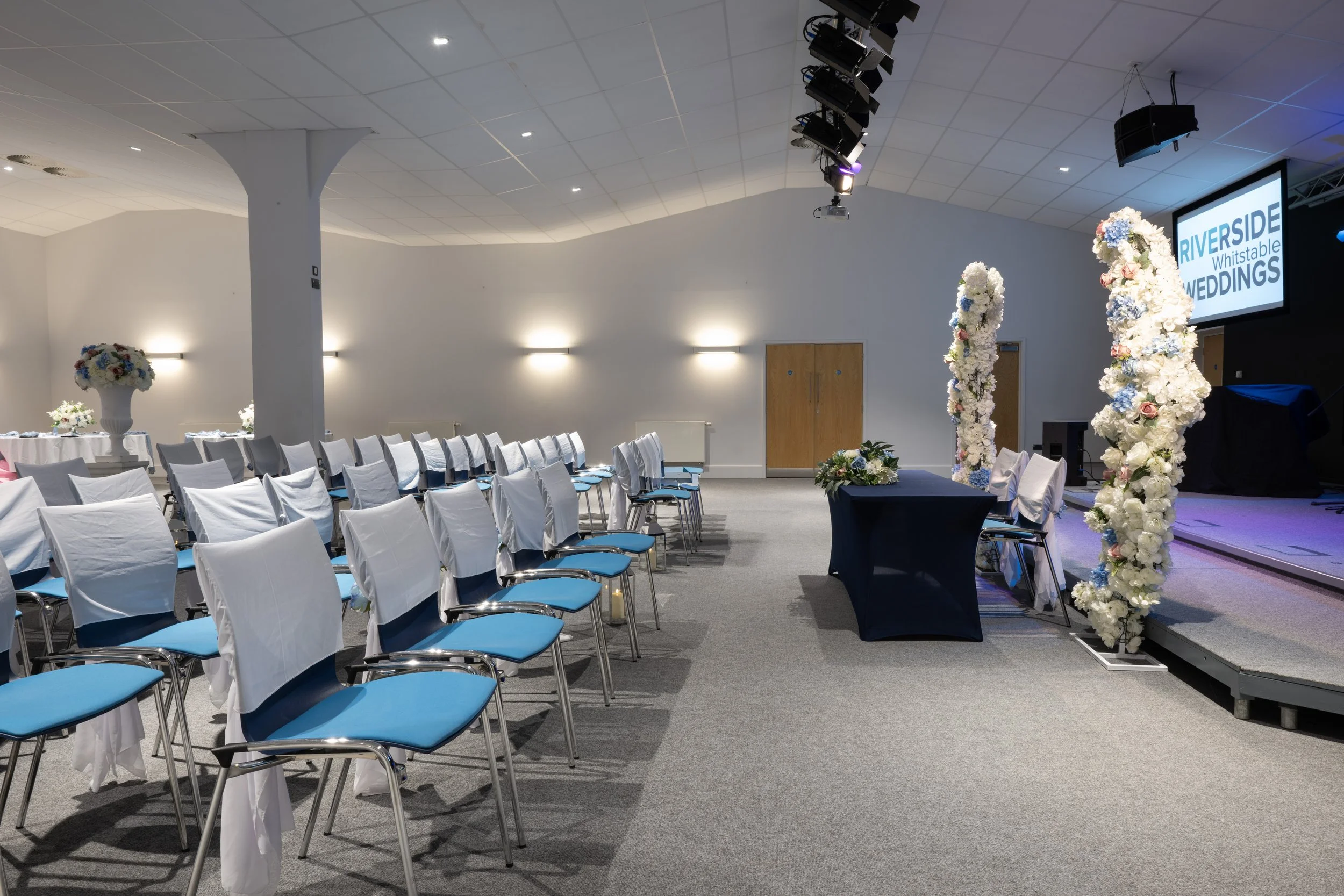 Empty wedding ceremony setup with rows of white chairs with blue cushions, decorated with white cloths, facing a stage with floral arches, a black table with floral arrangements, and a large screen displaying "RIVERSIDE Whistable WEDDINGS" in an even