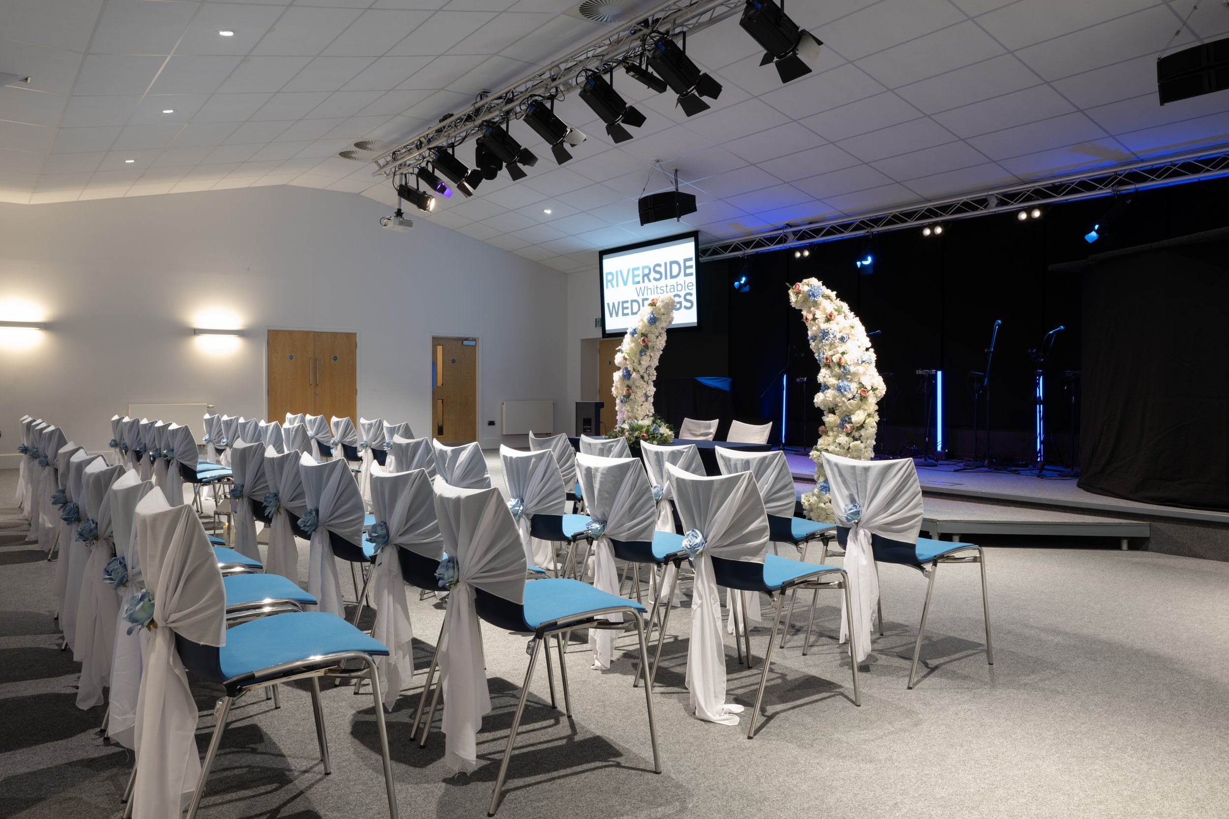An indoor venue prepared for a wedding ceremony with chairs covered in white cloth and decorated with blue and white flowers, arranged facing a stage with floral arch decorations and a screen displaying details of the event, which is the wedding of R