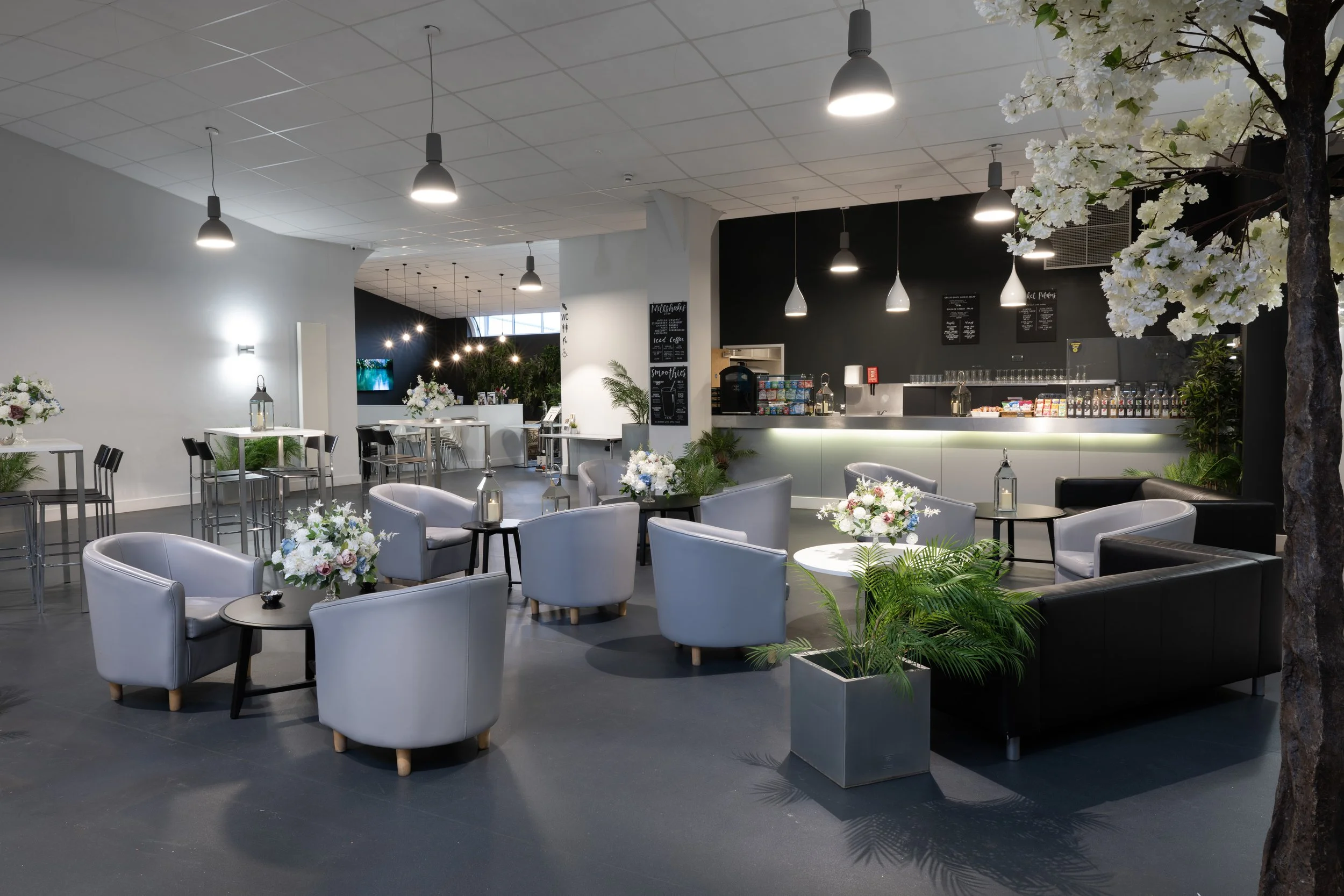 Modern indoor lounge area with gray armchairs, black sofas, round tables, flowers, a bar with drinks, and a black accent wall.