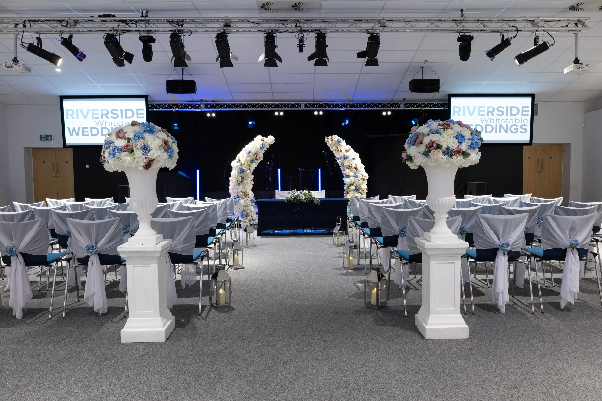 Indoor wedding venue decorated with large floral arrangements, arched floral structure at the front, chairs with white covers and blue ribbons, hanging lanterns, stage with microphones, and two large screens displaying "RIVERSIDE Whistable WEDDINGS".