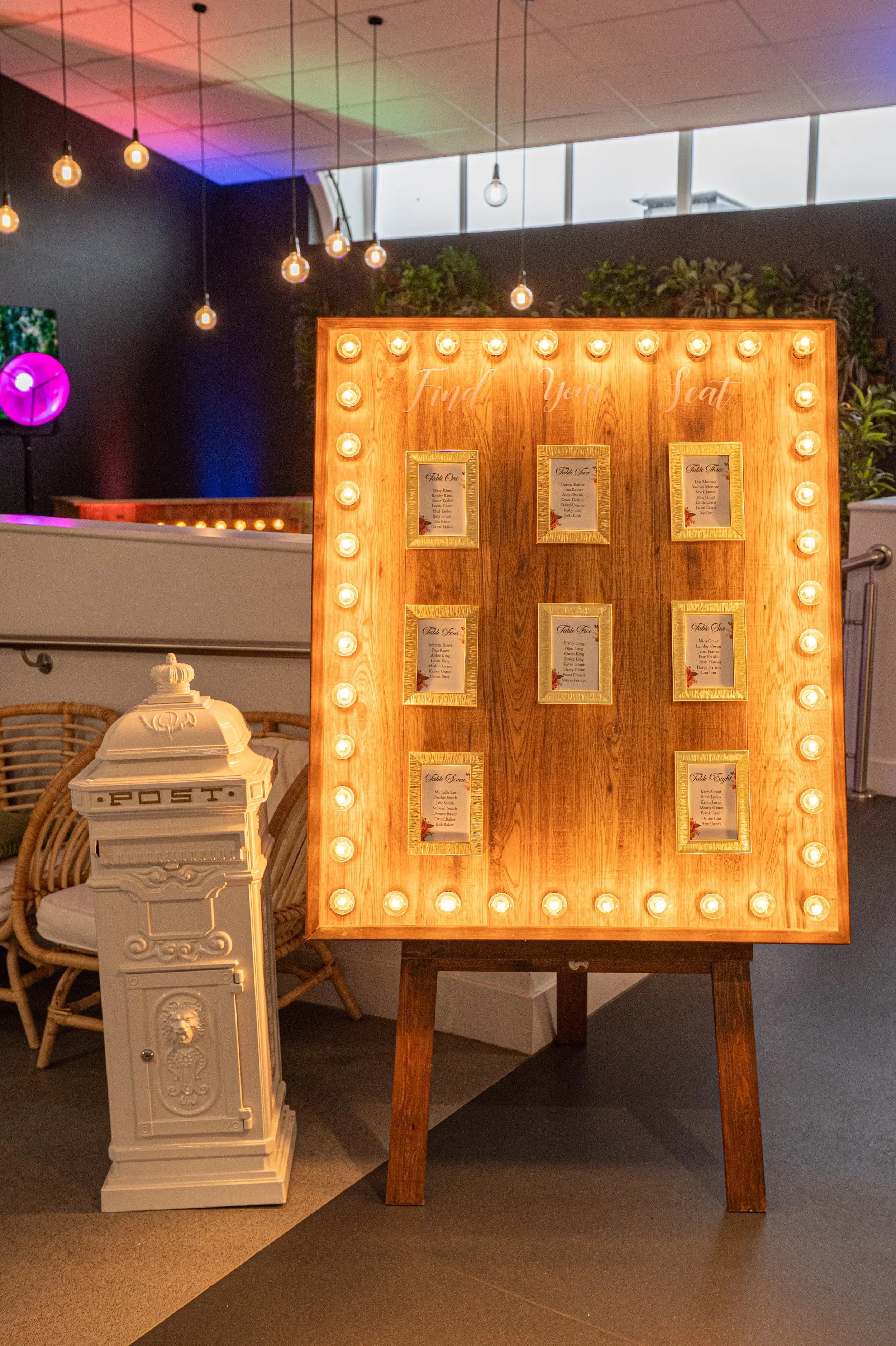 A wooden seating chart board with lightbulb lights surrounding it, and framed table numbers or names, in a decorated venue with a post box on the side.