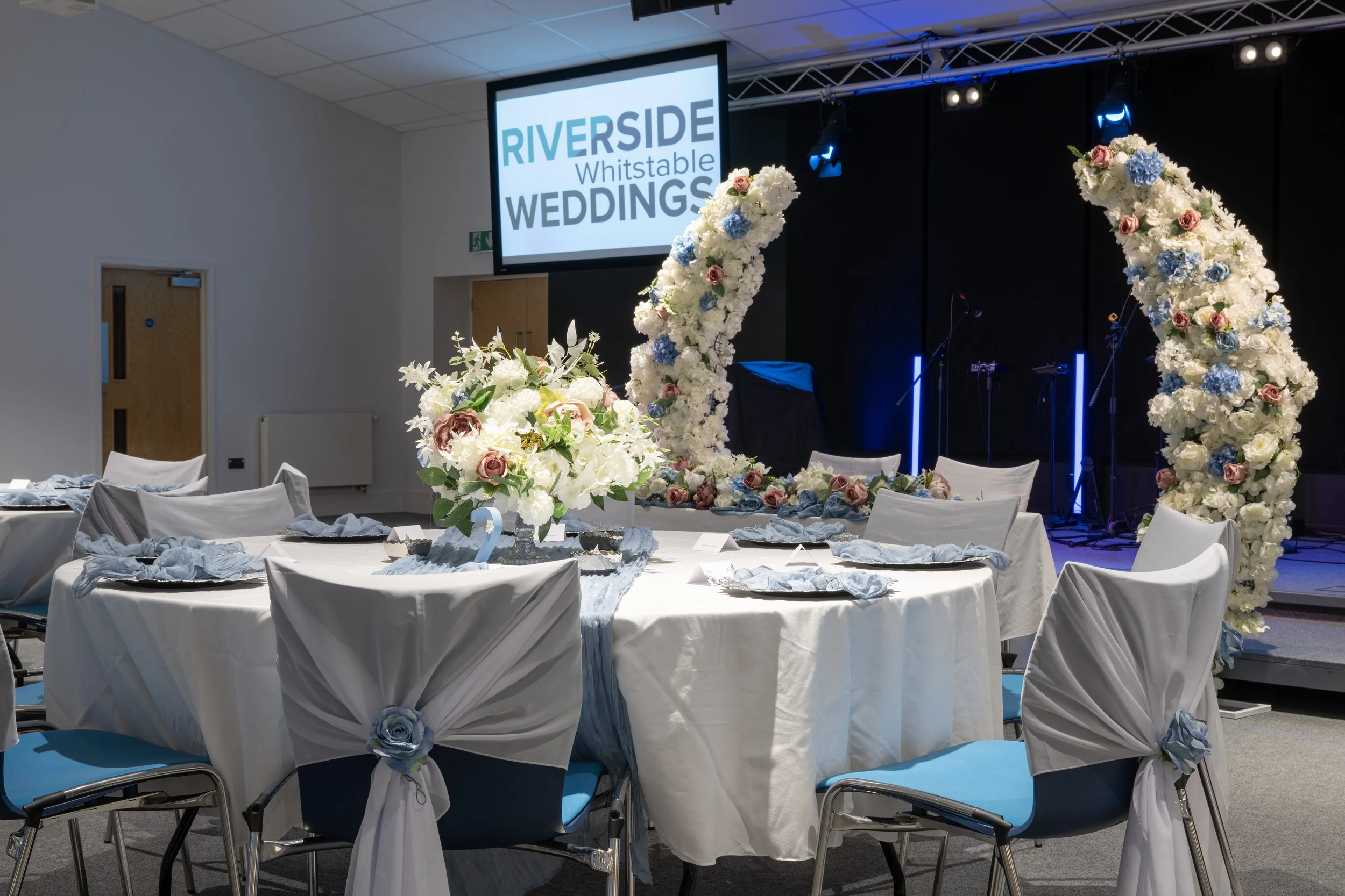 Wedding reception setup with decorated tables and chairs, floral arrangements, and a stage with a large screen displaying 'RIVERSIDE Whistable WEDDINGS'.