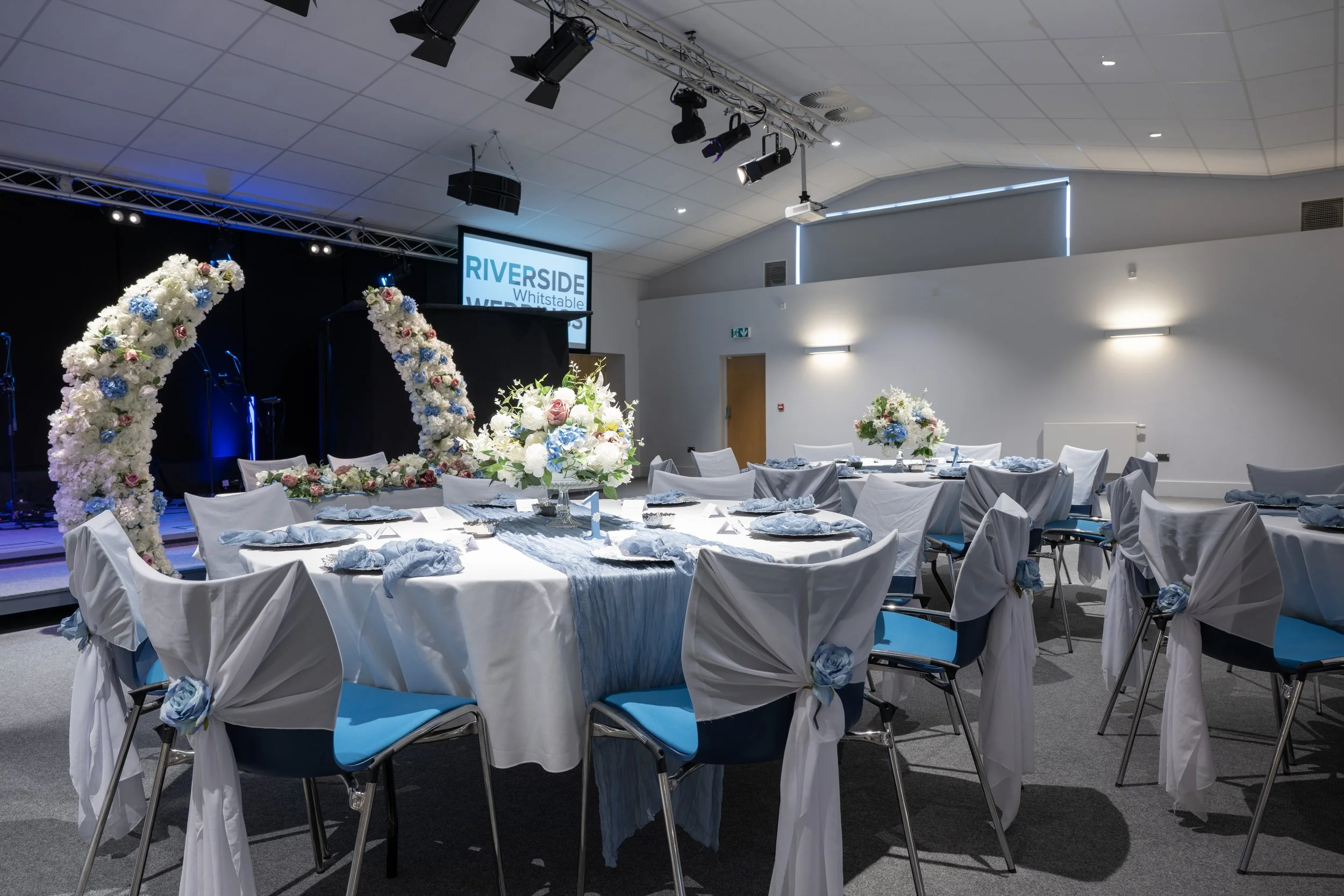 Indoor event space decorated with white and blue floral arrangements, round tables with white tablecloths and Blue chairs, stage with flower arches and a screen displaying event information.