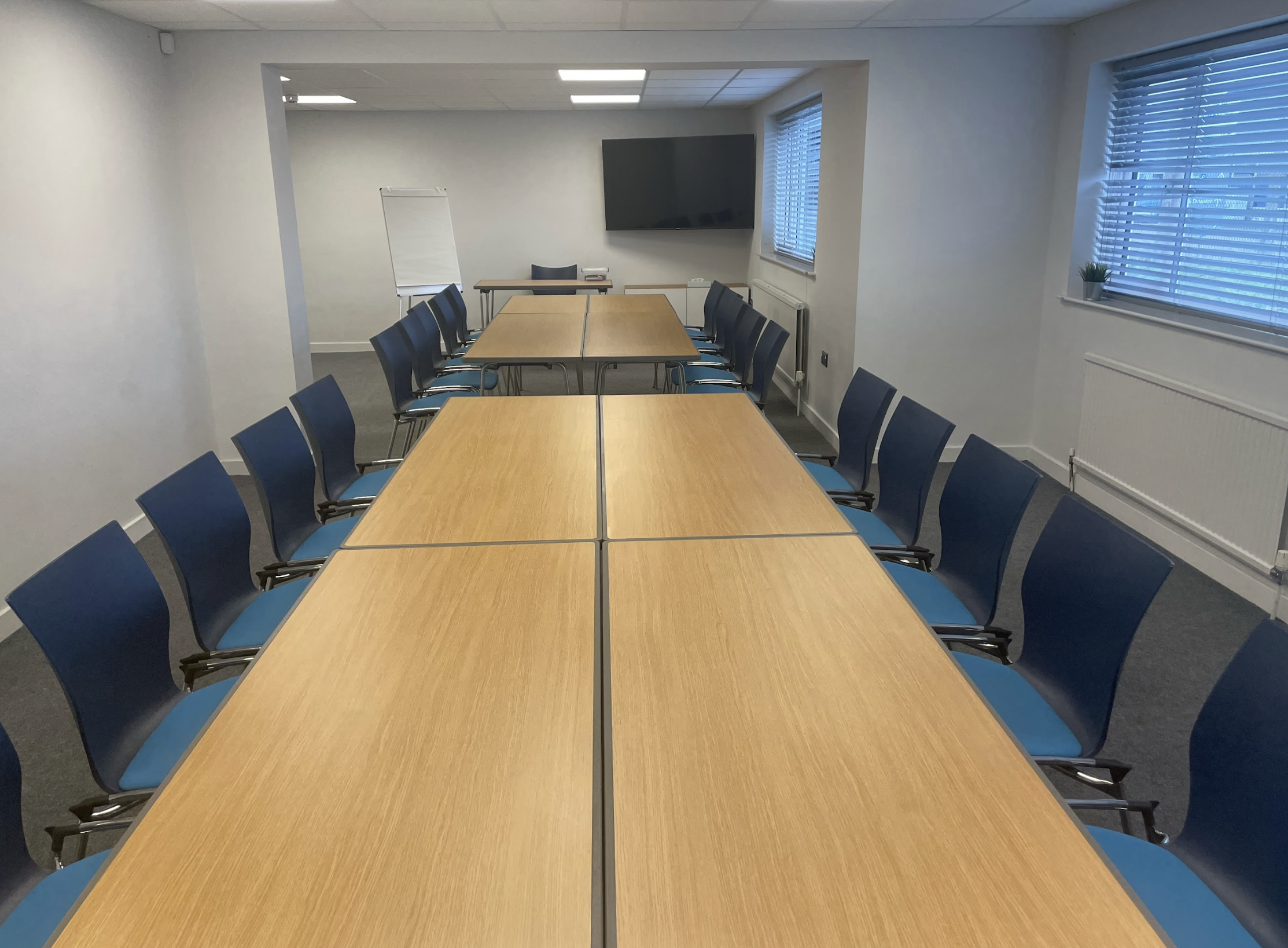 Empty conference room with wooden tables arranged in a U-shape and black chairs with blue cushions. There is a large flat-screen TV on the back wall, a whiteboard, and windows with blinds.