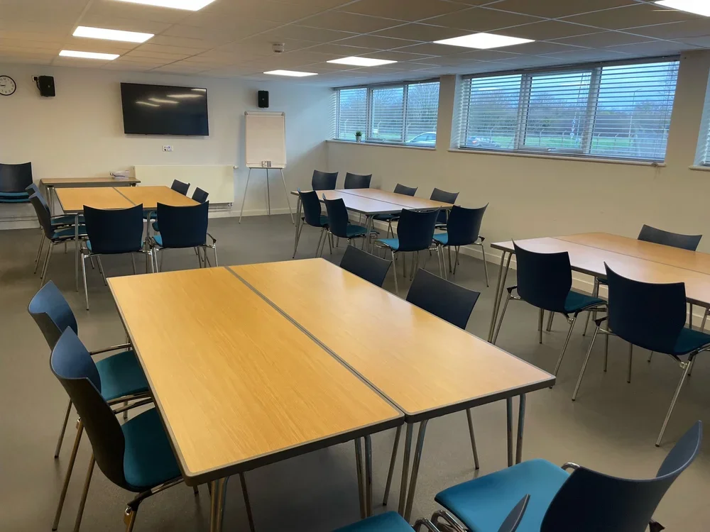 Empty conference room with wooden tables, blue and black chairs, a large flat-screen TV, a whiteboard, and windows with blinds.