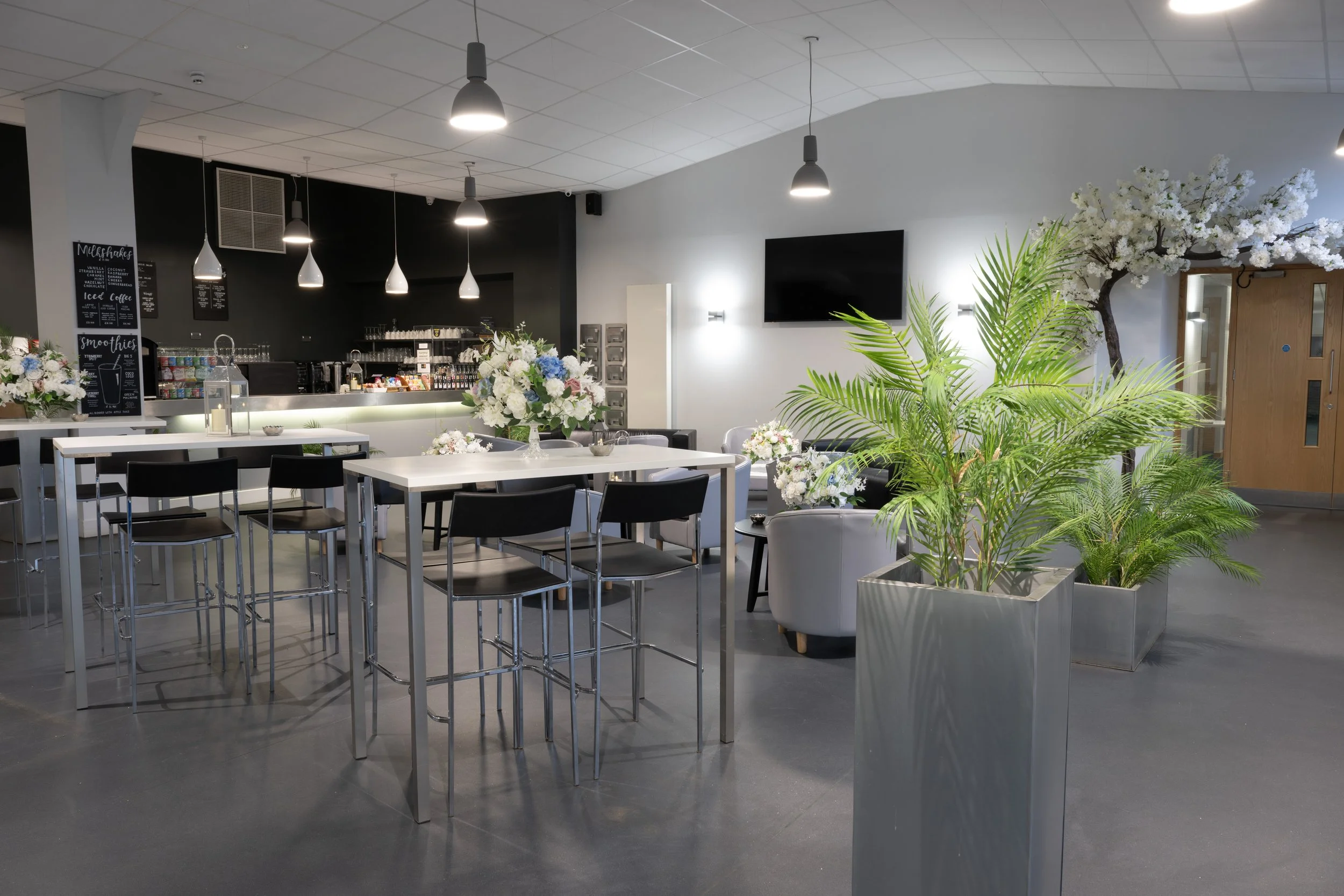 Modern indoor lounge area with white tables, black chairs, gray armchairs, and large planters with green plants and white flowers, decorated for a special event.