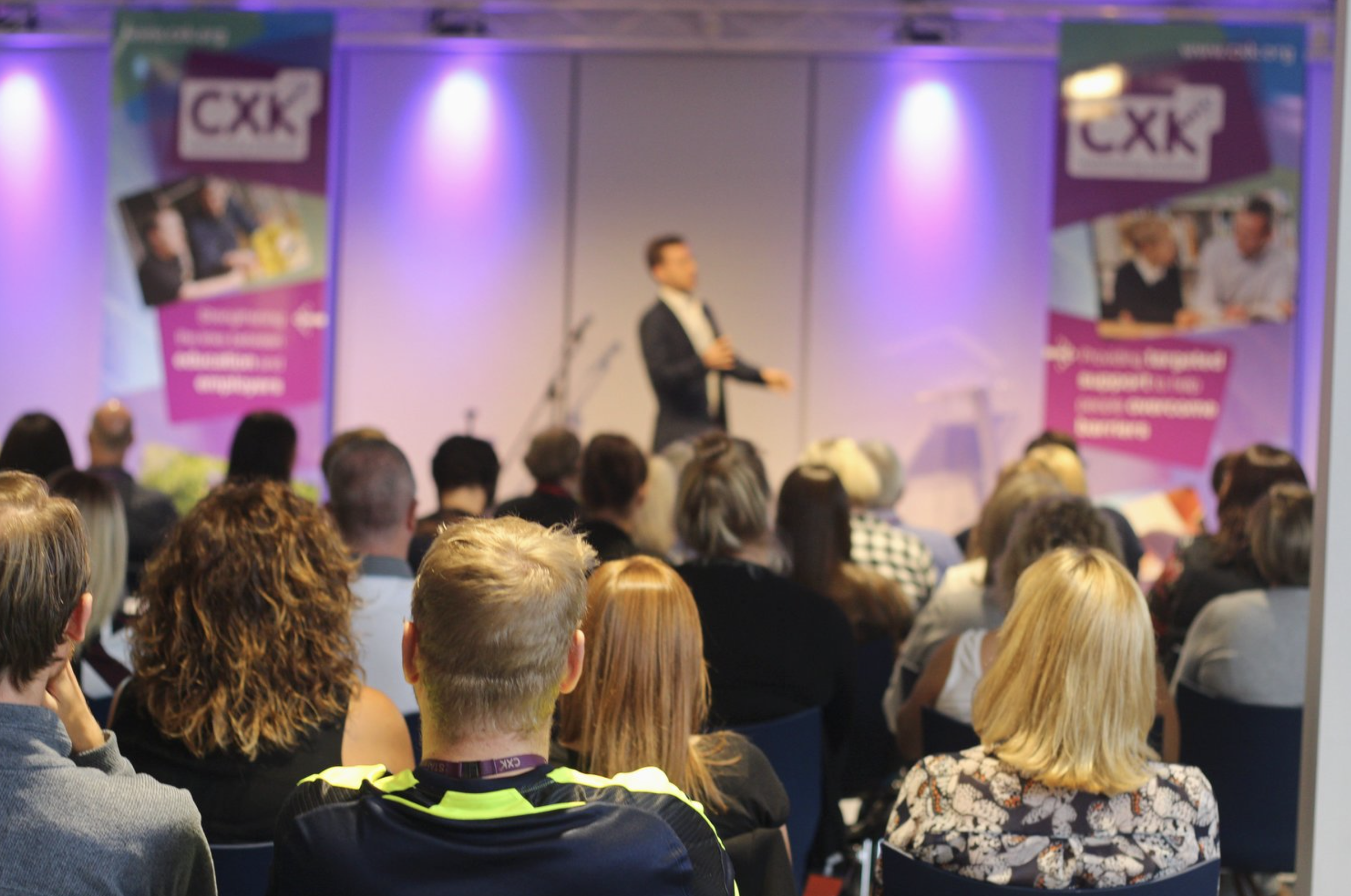 A man in a dark suit and white shirt giving a presentation on stage at a conference, with an audience of mostly adults watching. There are banners on either side of the stage that display the CXK logo and promotional images.