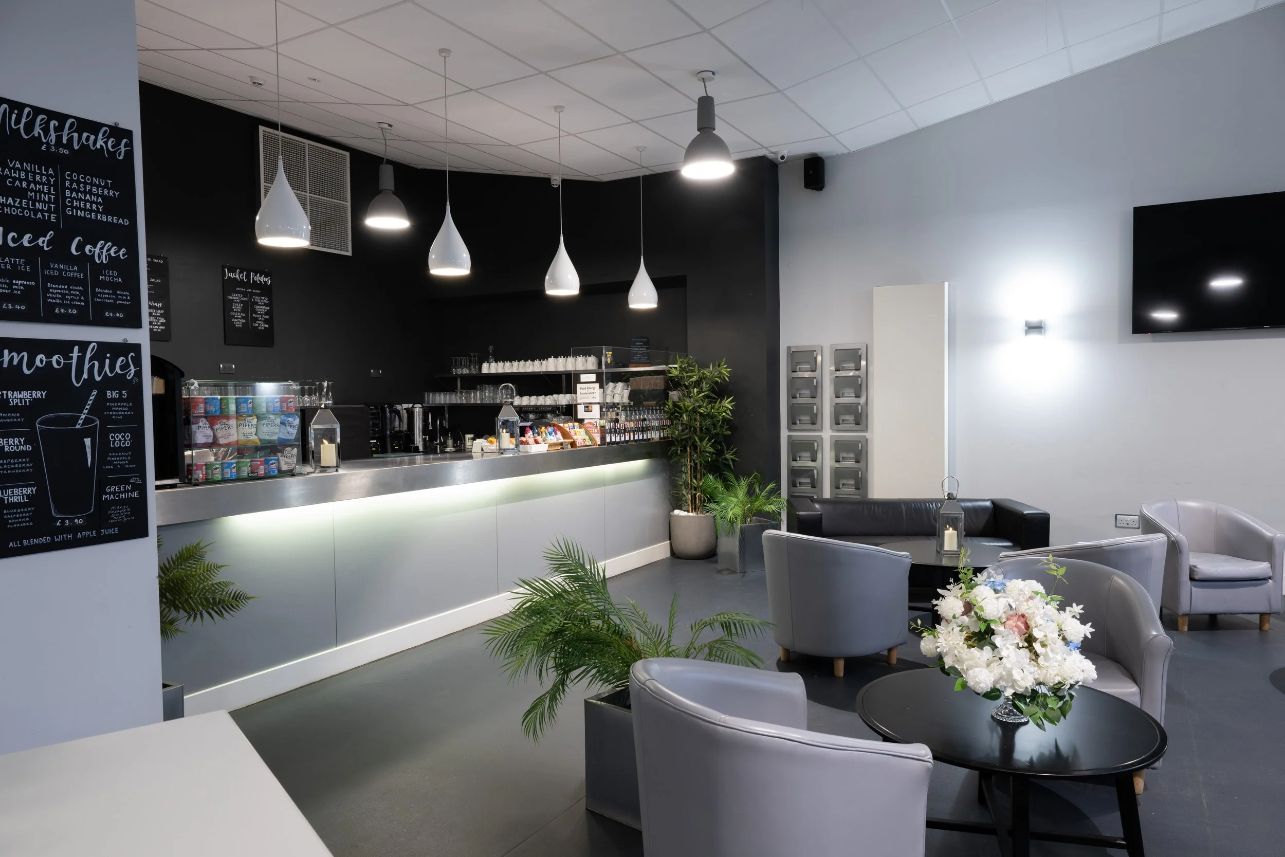 Modern coffee shop interior with a counter, menu boards, and seating area with gray chairs and a flower arrangement.