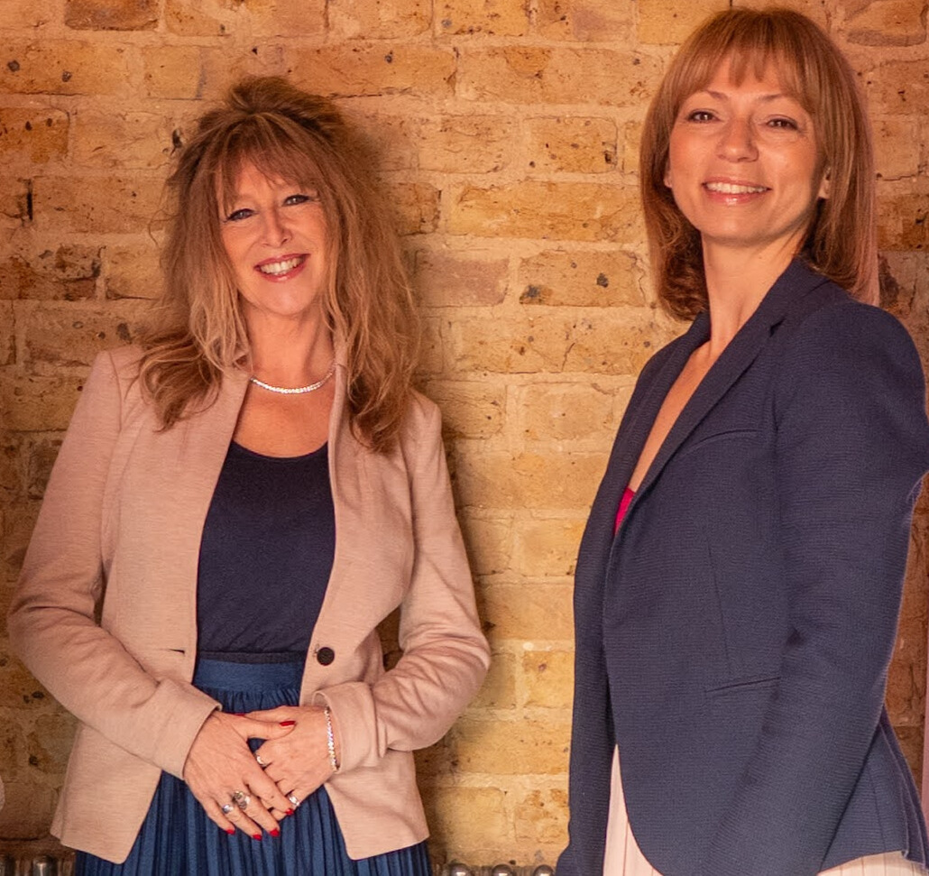 Two women smiling and standing in front of a brick wall. The woman on the left has long, wavy, reddish-blonde hair, and is wearing a beige blazer over a dark top, with jewelry. The woman on the right has straight, shoulder-length, light brown hair, and is wearing a navy blazer over a top.