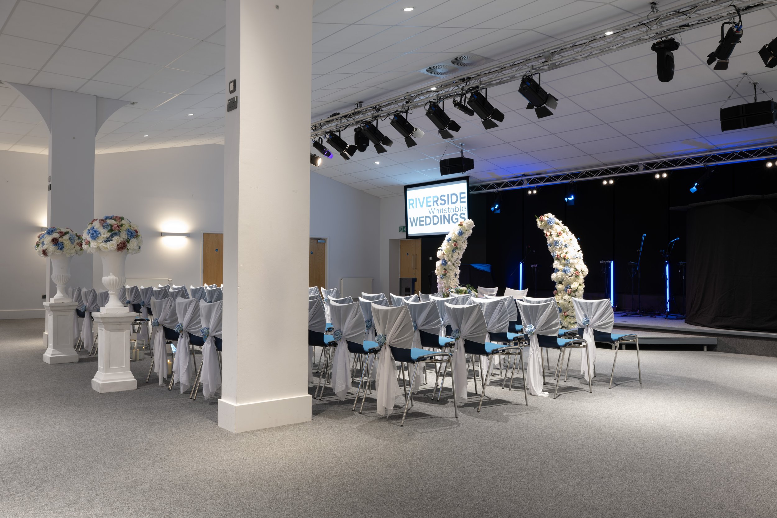 Wedding ceremony setup with chairs decorated with white covers and blue sashes, floral arrangements on pedestals, and a stage with a large screen displaying 'RIVERSIDE WEDDINGS' in Whitstable. The stage includes floral arches and a musical setup with