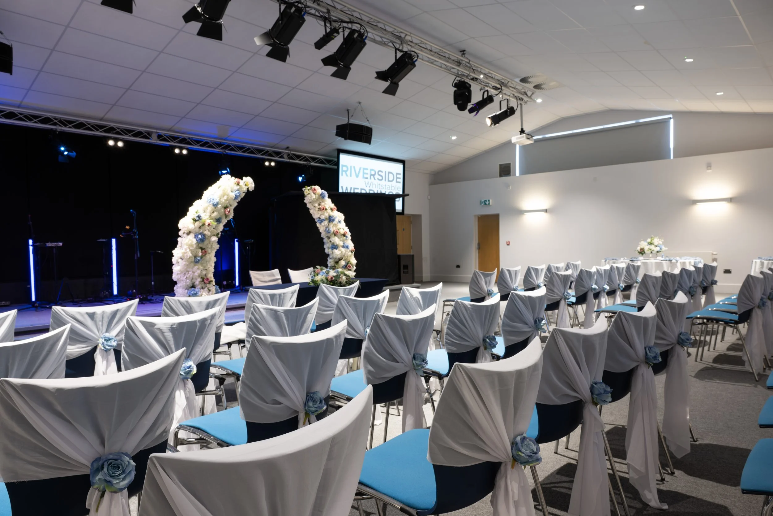 An indoor event space set up for a wedding, with chairs decorated with white fabric and blue flowers, facing a stage with floral arches and musical instruments, and a screen displaying the words 'RIVERSIDE Whitstable WEDDING.'