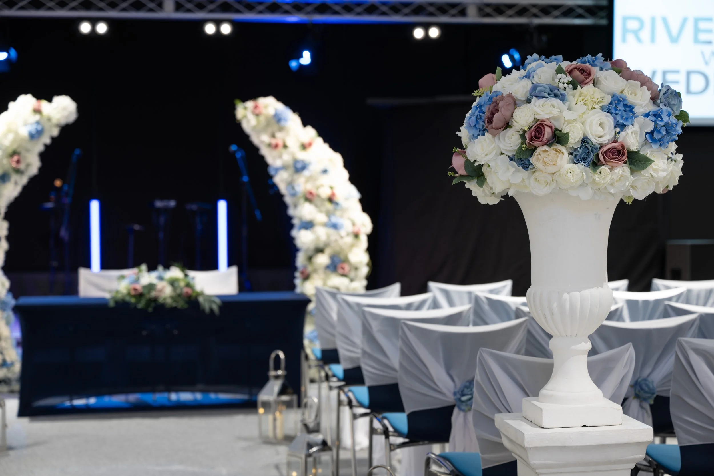 Elegant event setup with a large white floral arrangement and chairs draped in white fabric, decorated with blue and pink flowers, overlooking a stage with arc-shaped floral arches.