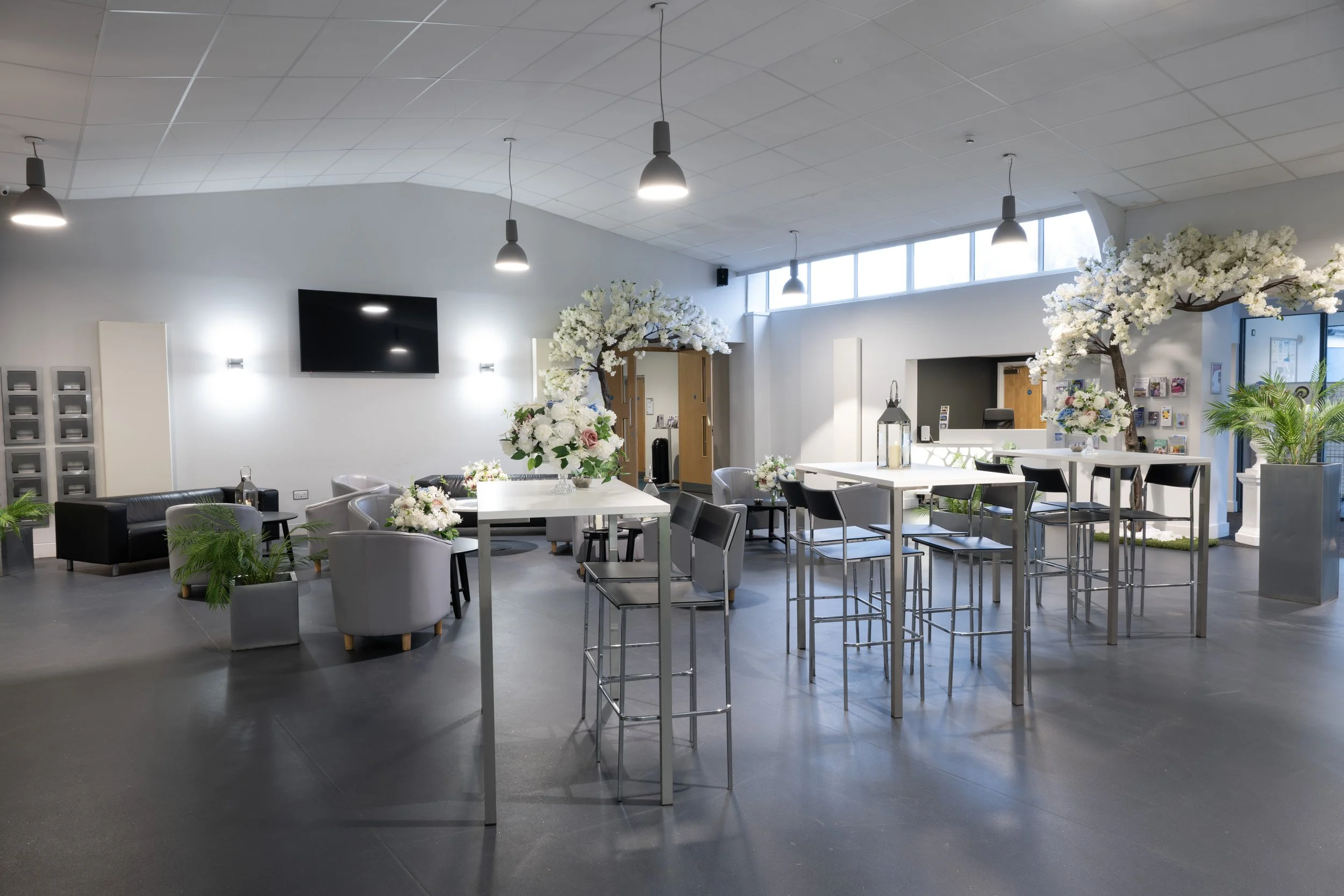 Interior of a modern event space decorated with floral arrangements and greenery for a celebration, featuring tables, chairs, and soft lighting.