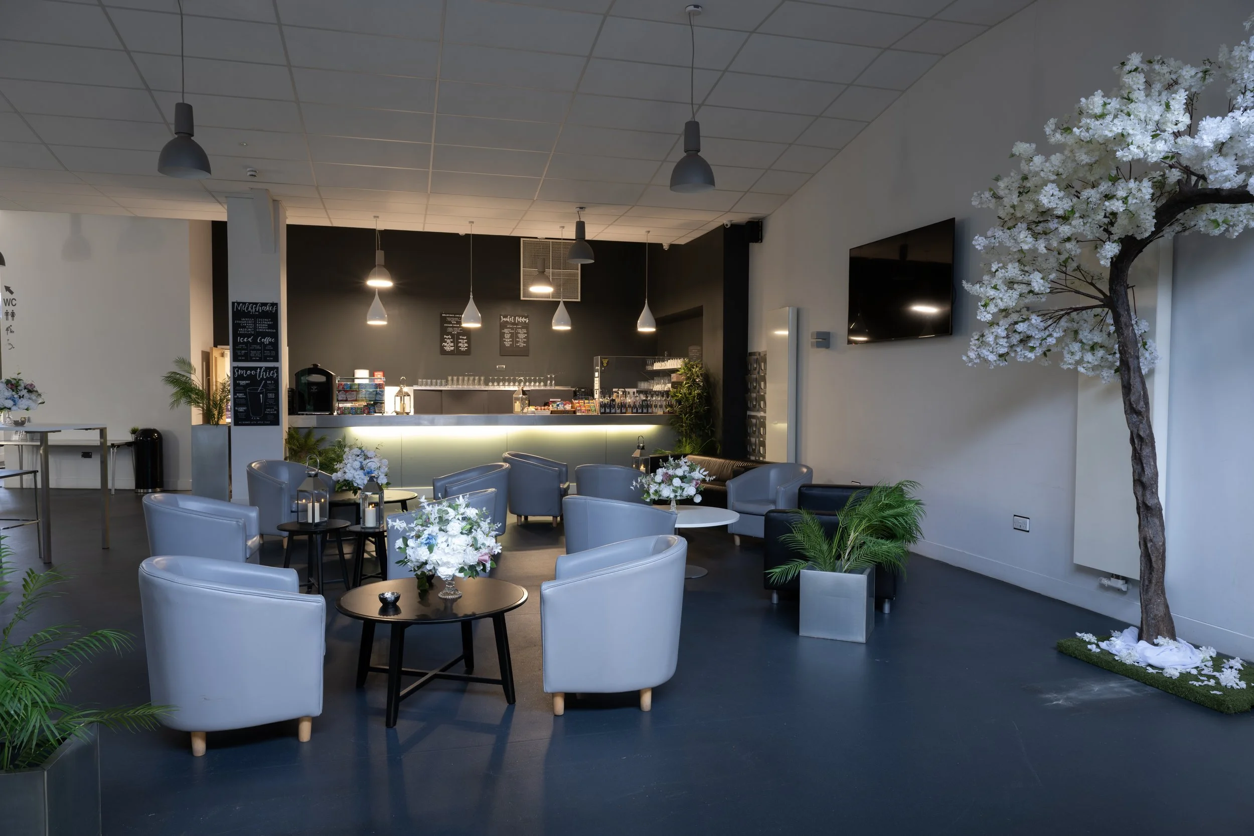 Interior of a modern cafe with white and grey furniture, floral arrangements, potted plants, a bar counter in the background, and a large tree decoration on the right side.