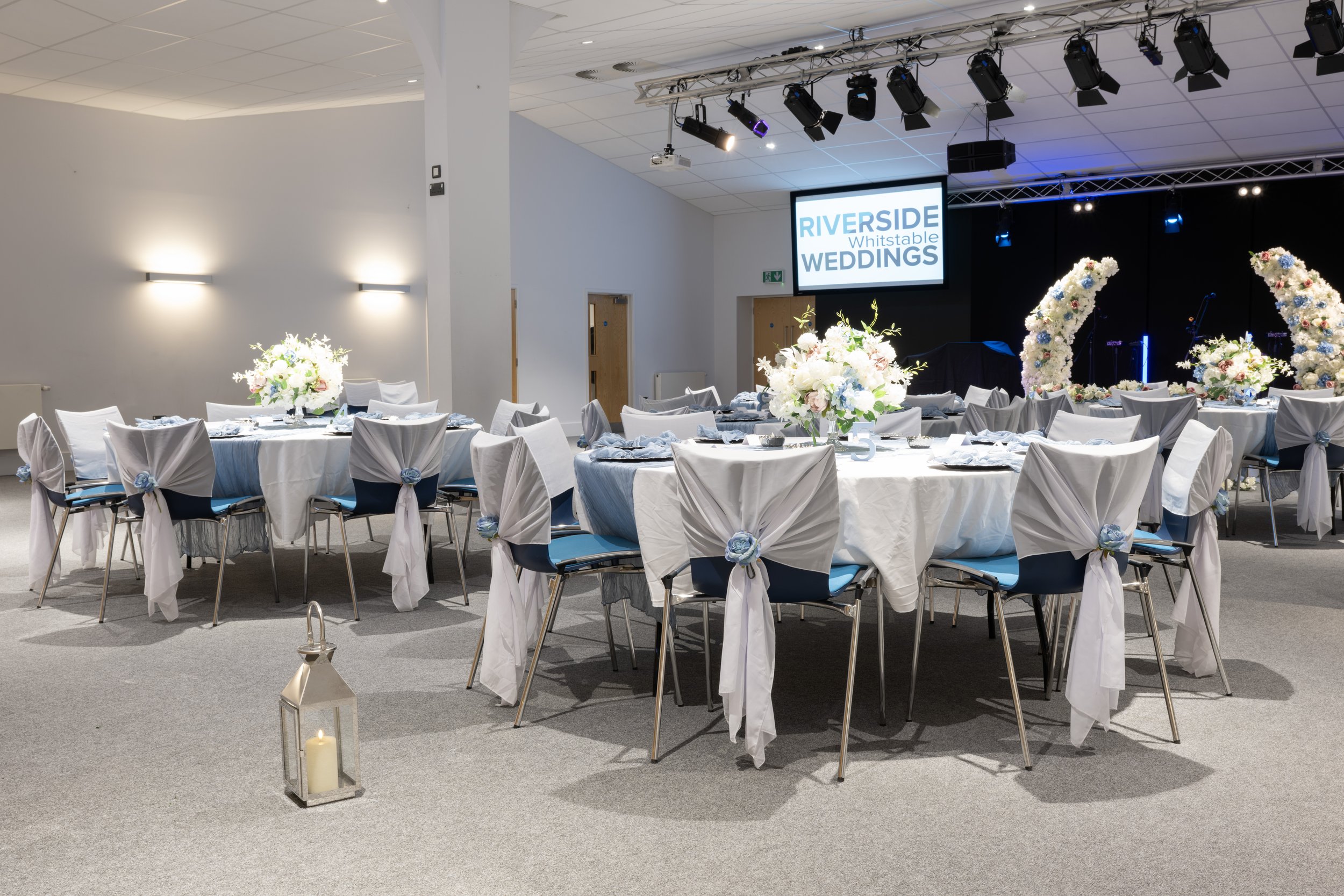 Decorated wedding reception hall with round tables covered in white tablecloths, chairs with white fabric drapes tied with blue flowers, floral centerpieces, and a stage with a large sign that reads 'RIVERSIDE Whistable WEDDINGS' and flower arches.