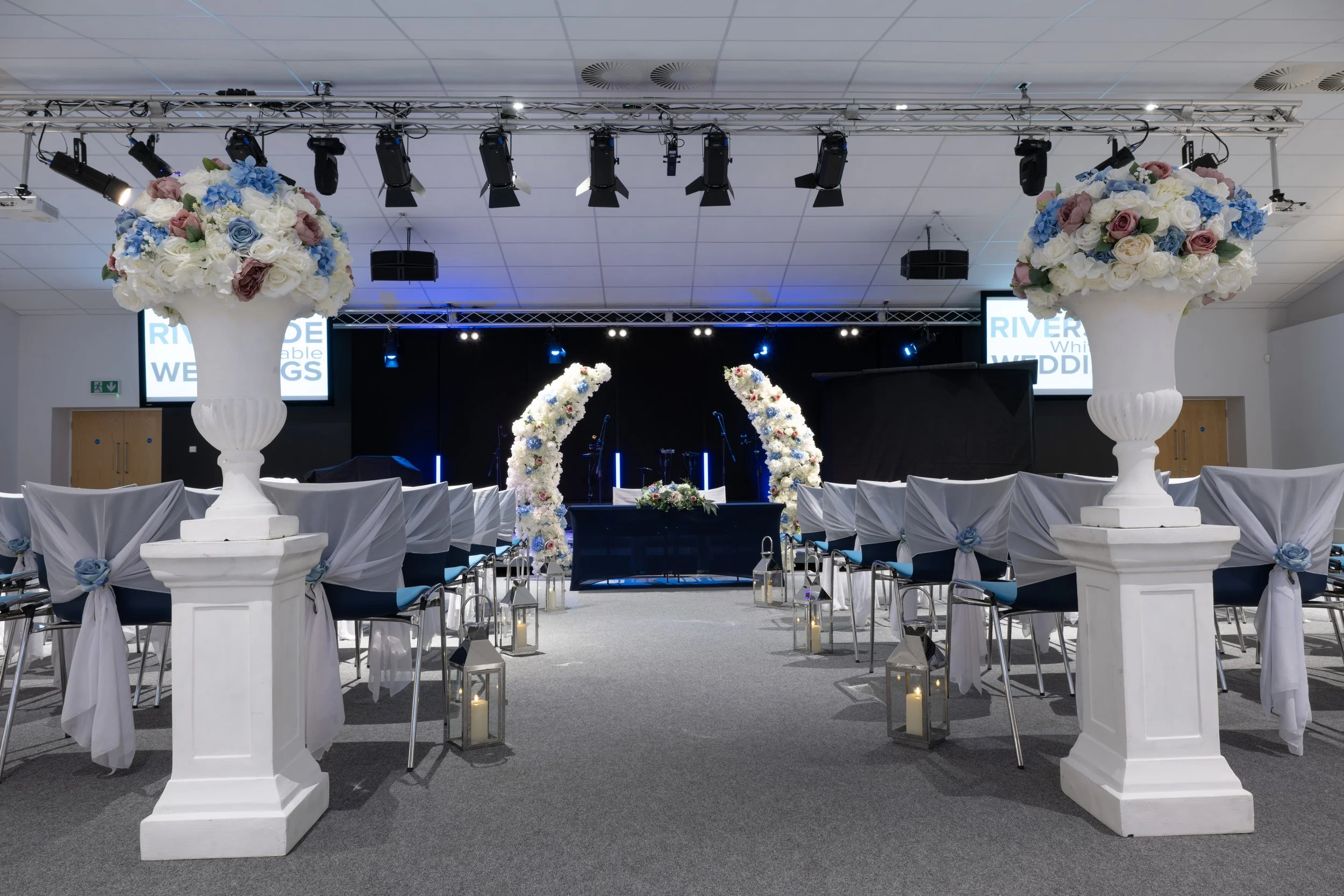 Indoor wedding ceremony setup with white chairs decorated with white cloth and blue ribbons, large floral arrangements of roses and hydrangeas in white urns on pedestals, floral arches of white and colorful flowers in the center, lanterns with candle