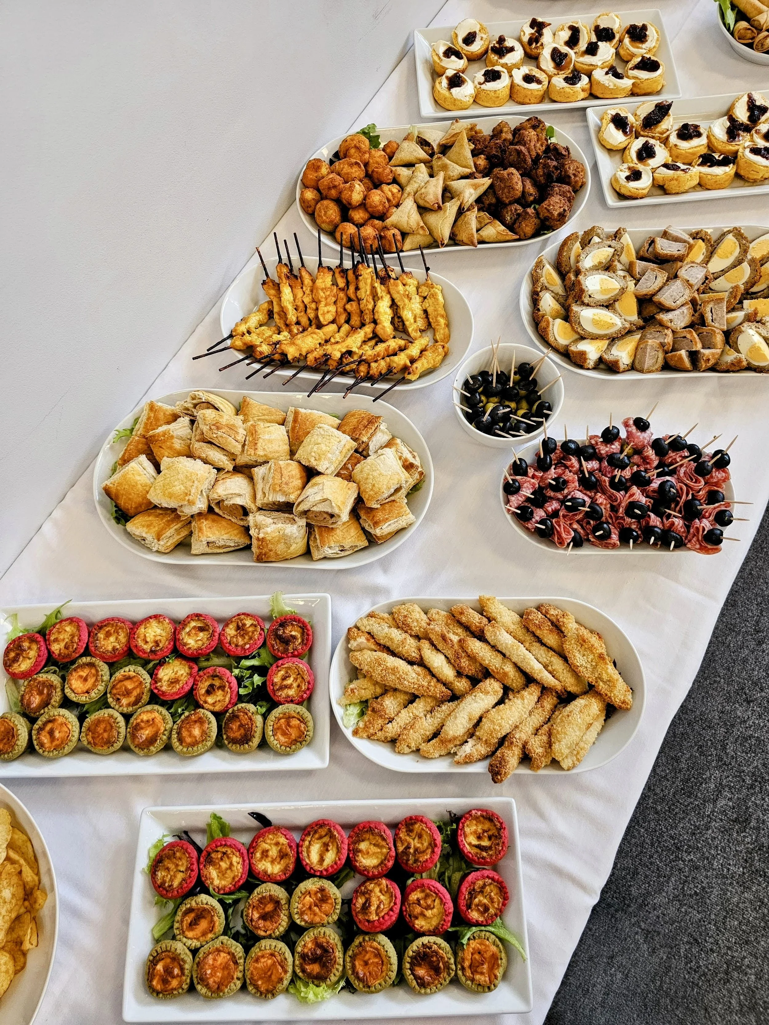 A buffet table filled with a variety of appetizers and finger foods, including mini quiches, fried chicken skewers, pastries, fruit slices, and small sandwiches, arranged on white serving platters.