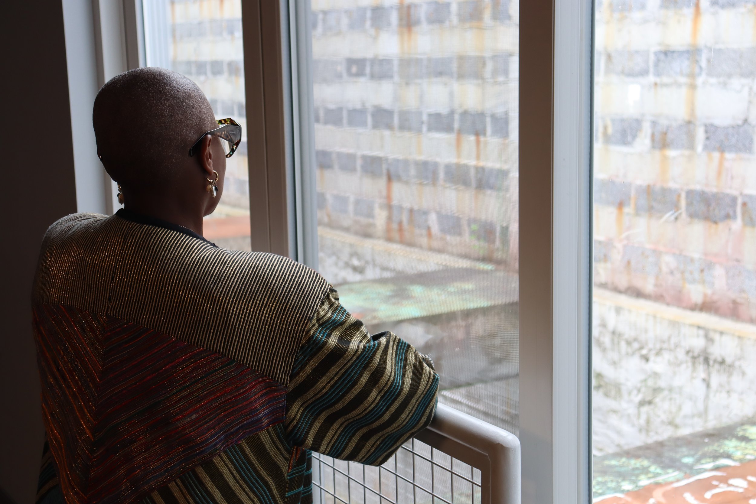 A person with a short hairstyle, wearing glasses and a striped top, looking out of a window at a brick wall outside.
