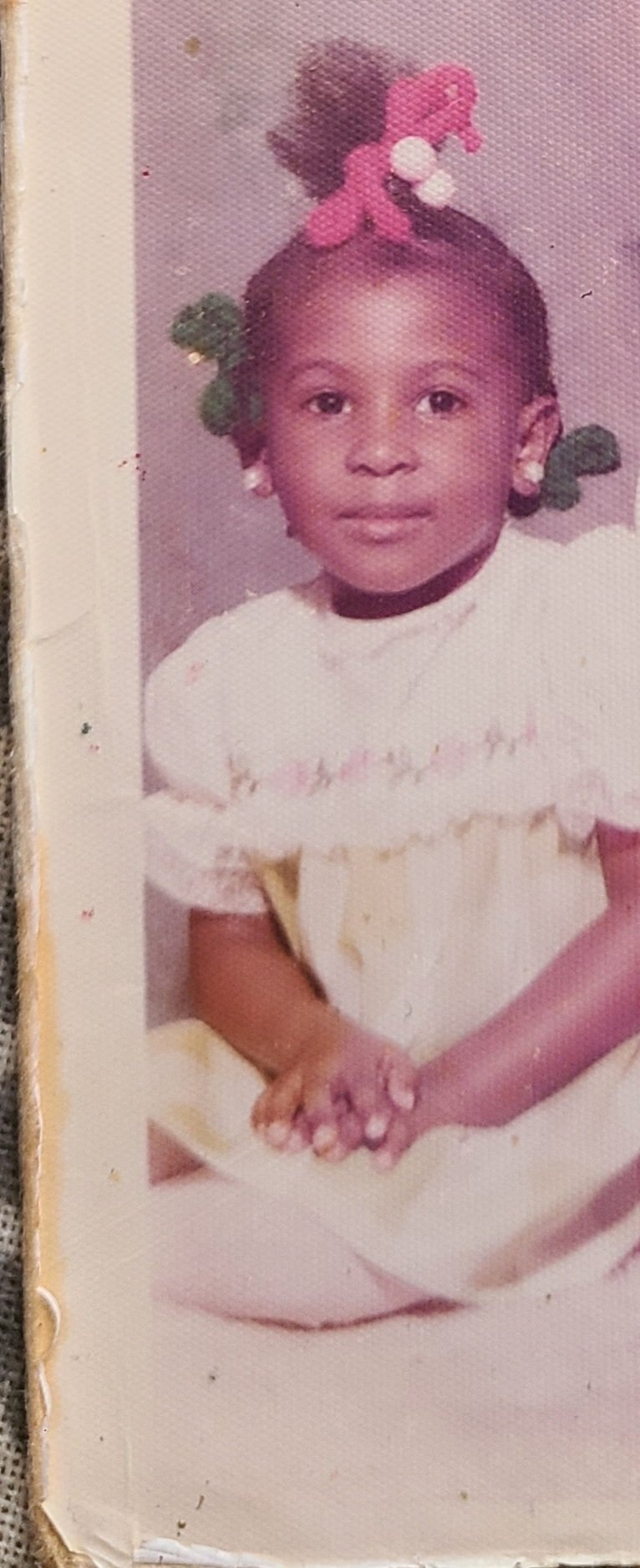 A young girl with braided hair decorated with pink and green bows, sitting with her hands clasped in her lap, wearing a cream-colored dress with ruffles.