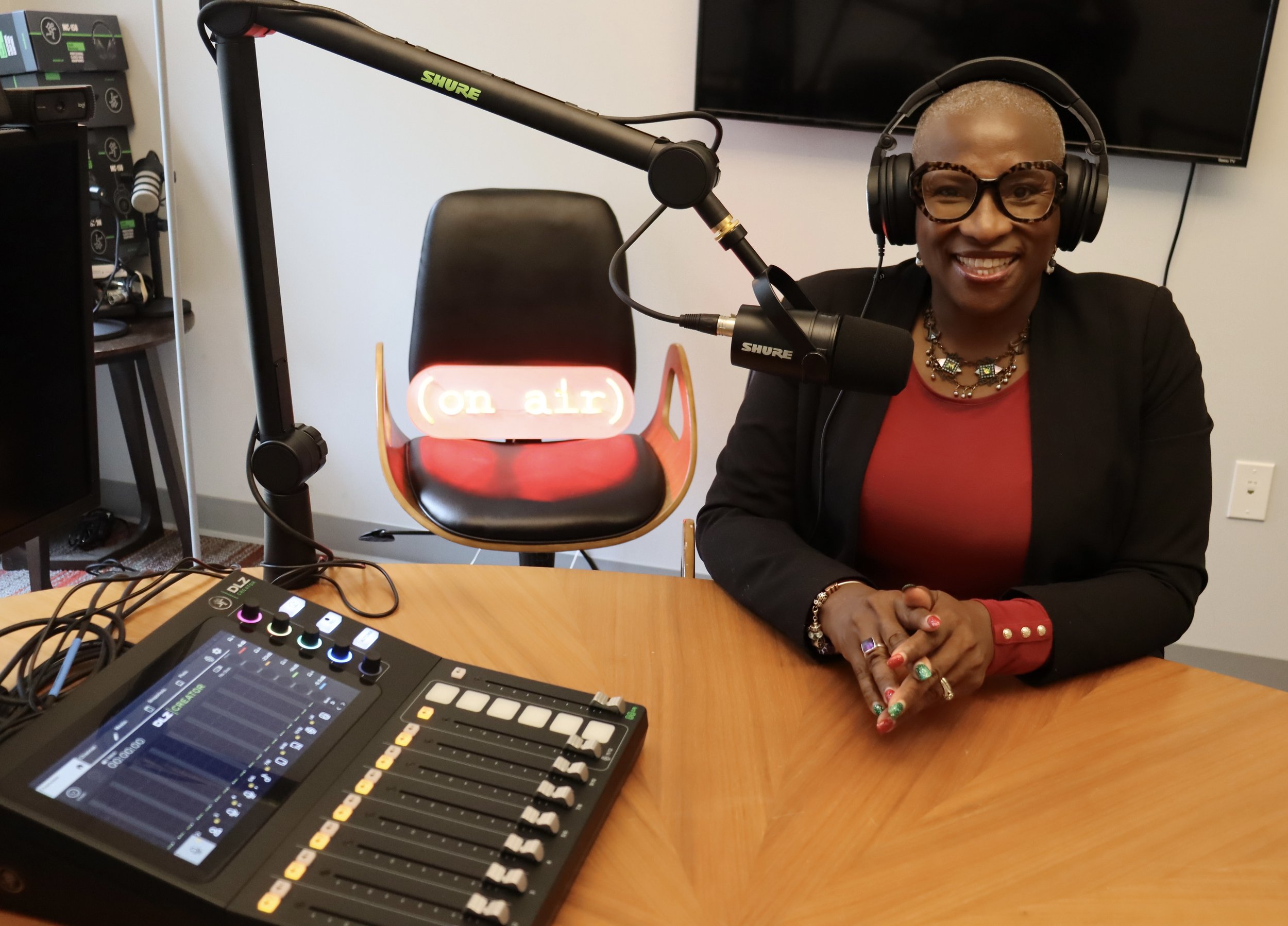 A woman with short gray hair, wearing glasses and a black blazer over a red top, sitting at a radio or podcast studio. She is smiling and wearing a large headset, with a microphone in front of her. On the table, there is audio mixing equipment and a chair with a red "On Air" sign.