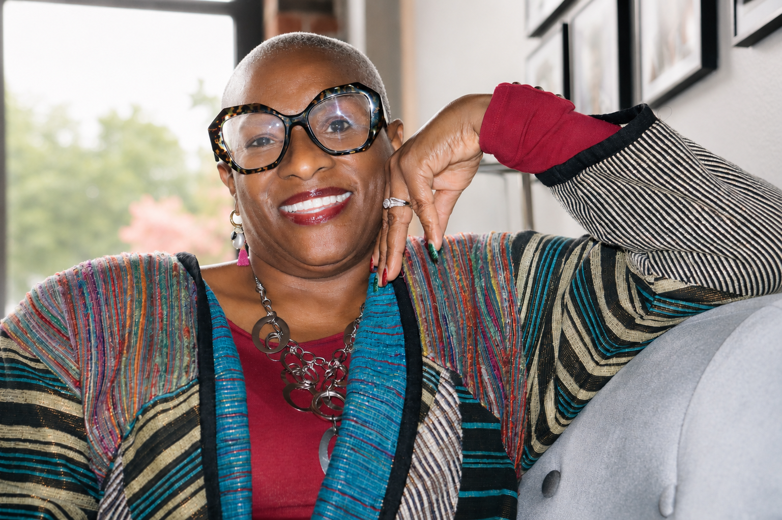 Close-up of a smiling woman with short, platinum blonde hair, wearing glasses, colorful striped jacket, red shirt, and jewelry, sitting on a light-colored couch indoors with a window and trees in the background.
